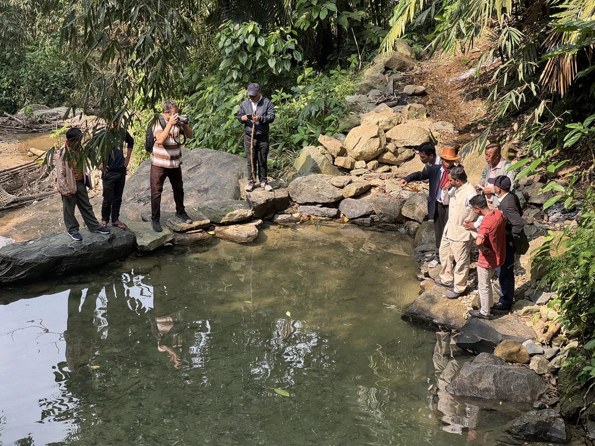 A group of people stand near a body of water. One man is filming the rest of the group.