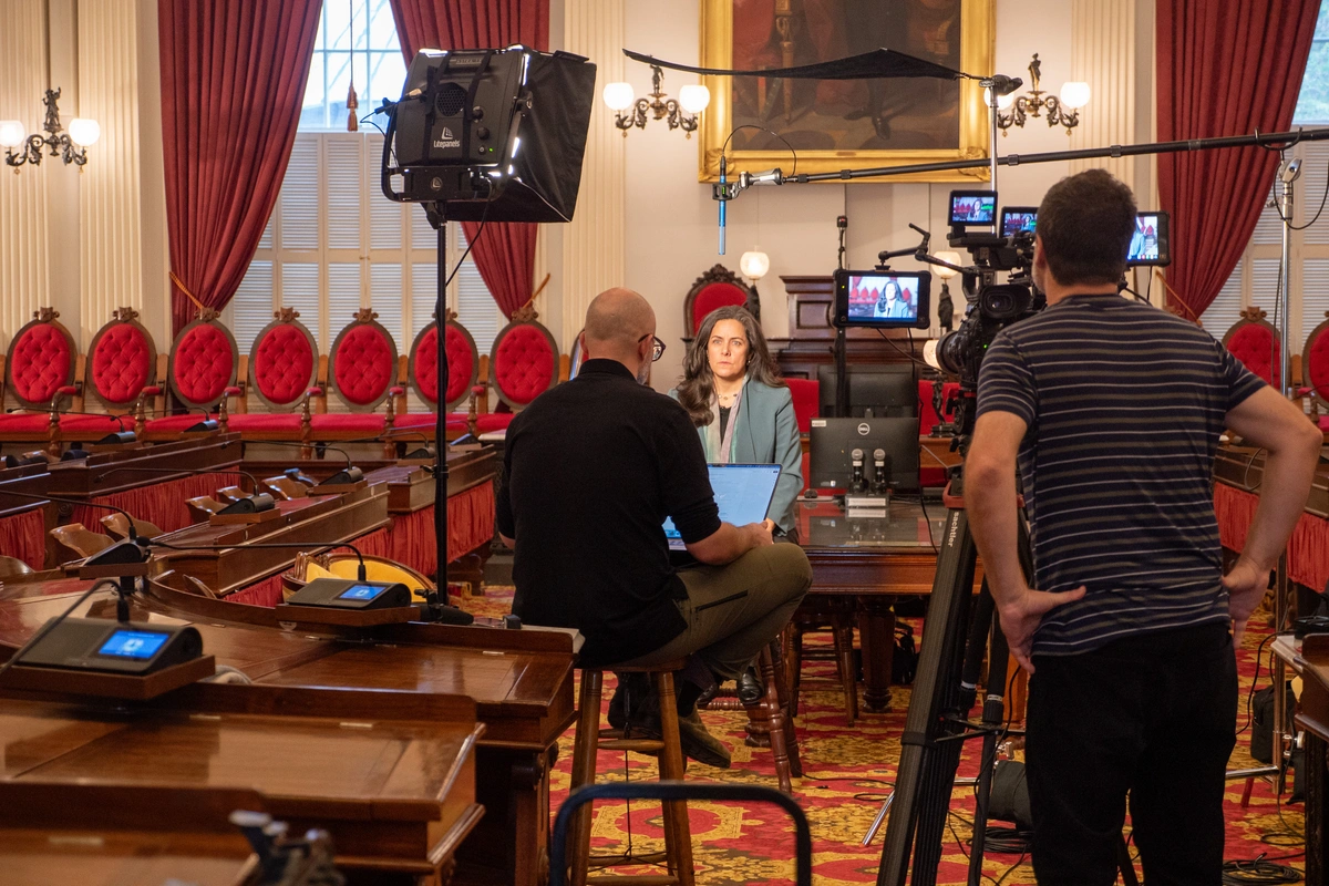 A woman in a blue blazer sits in front of a camera crew being interviewed.