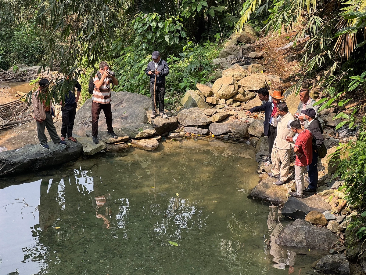 A group of people stand near a small body of water. One man is holding a camera and filming the rest.