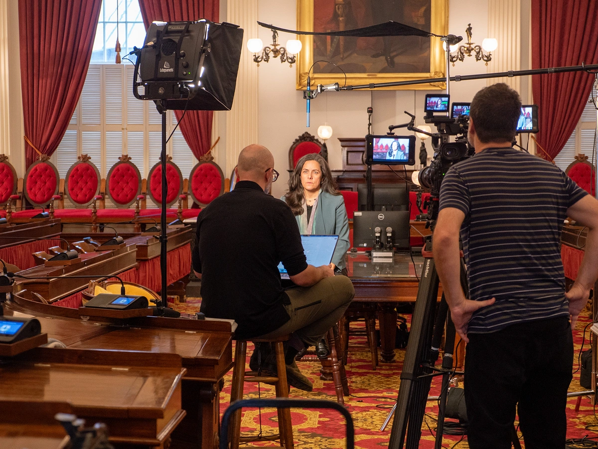 A woman in a blue balzer sits in front of a camera crew being filmed.