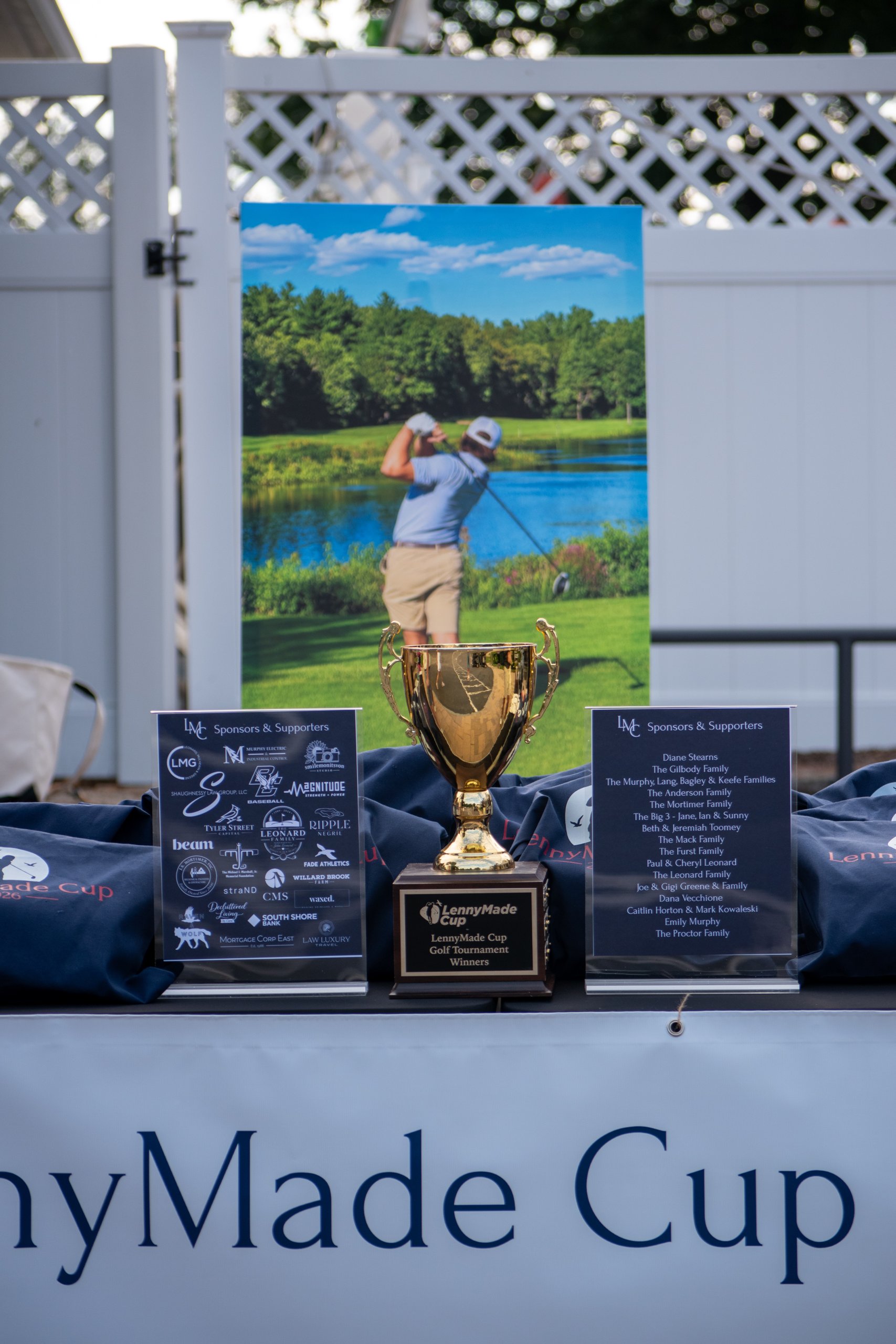 LennyMade Cup gold trophy on a table between sponsor and supporter signs, with navy LennyMade Cup gift bags and a golfer banner behind.