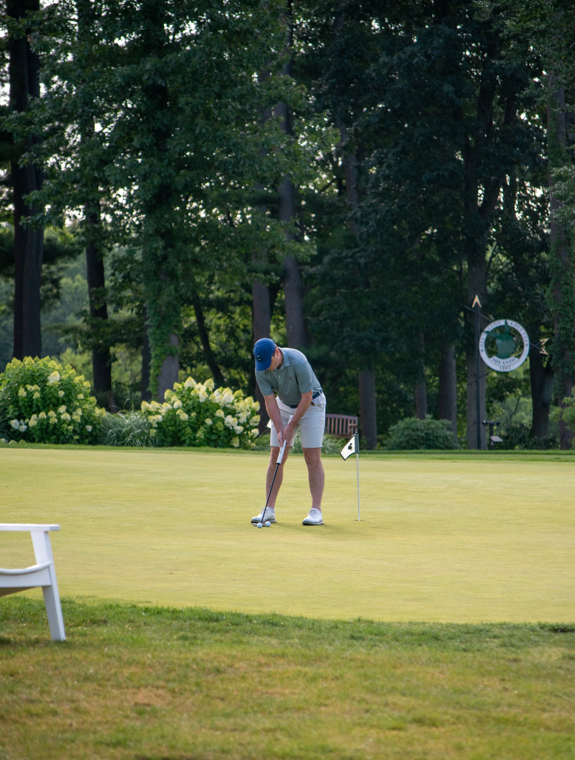 Golfer in a blue cap and gray polo putting on the green beside the flag, with a Sound View Golf Club sign among the trees behind.