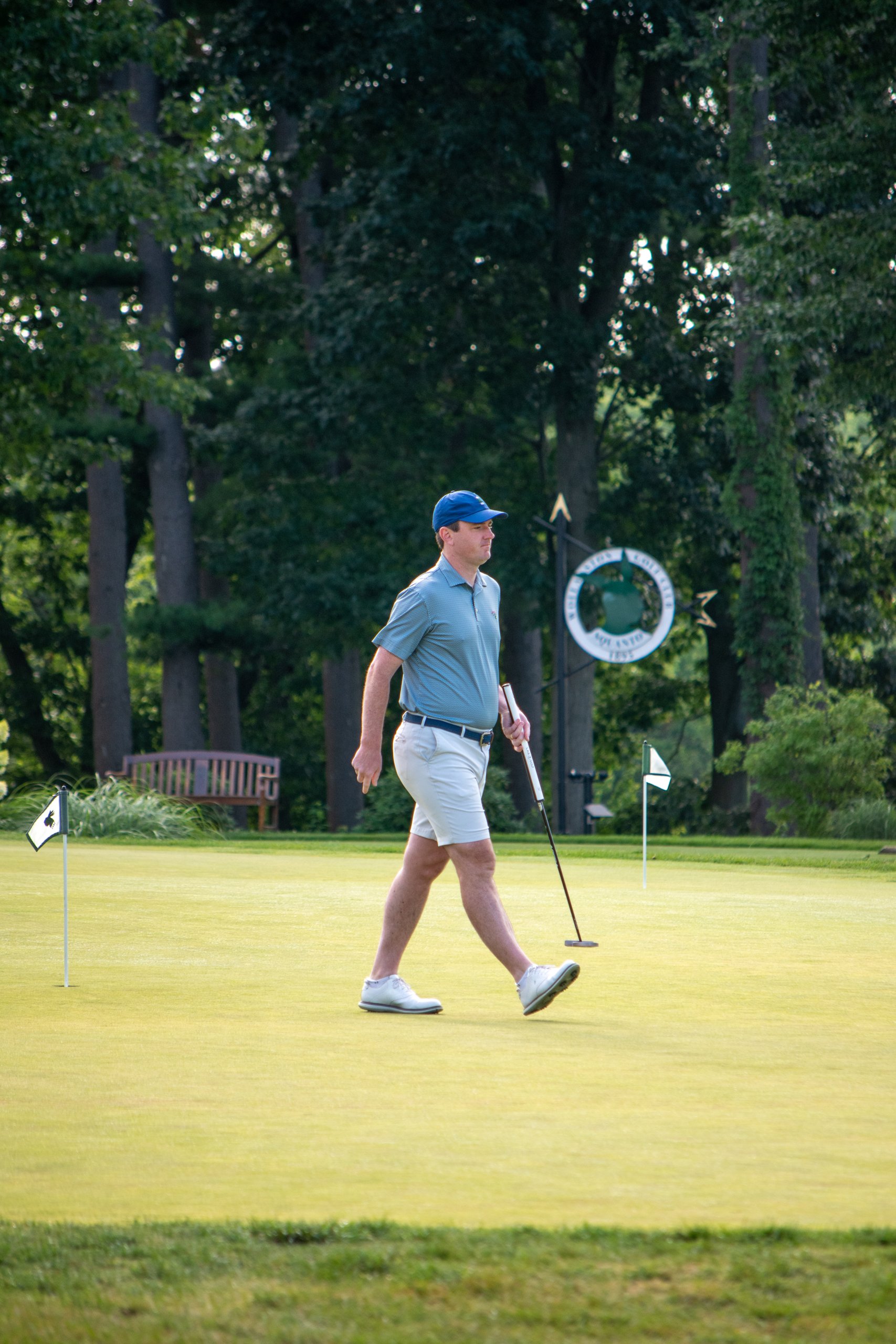 Golfer in a blue cap and gray polo walking across the putting green with a putter, a practice flag on each side and the Sound View Golf Club sign in the trees behind.