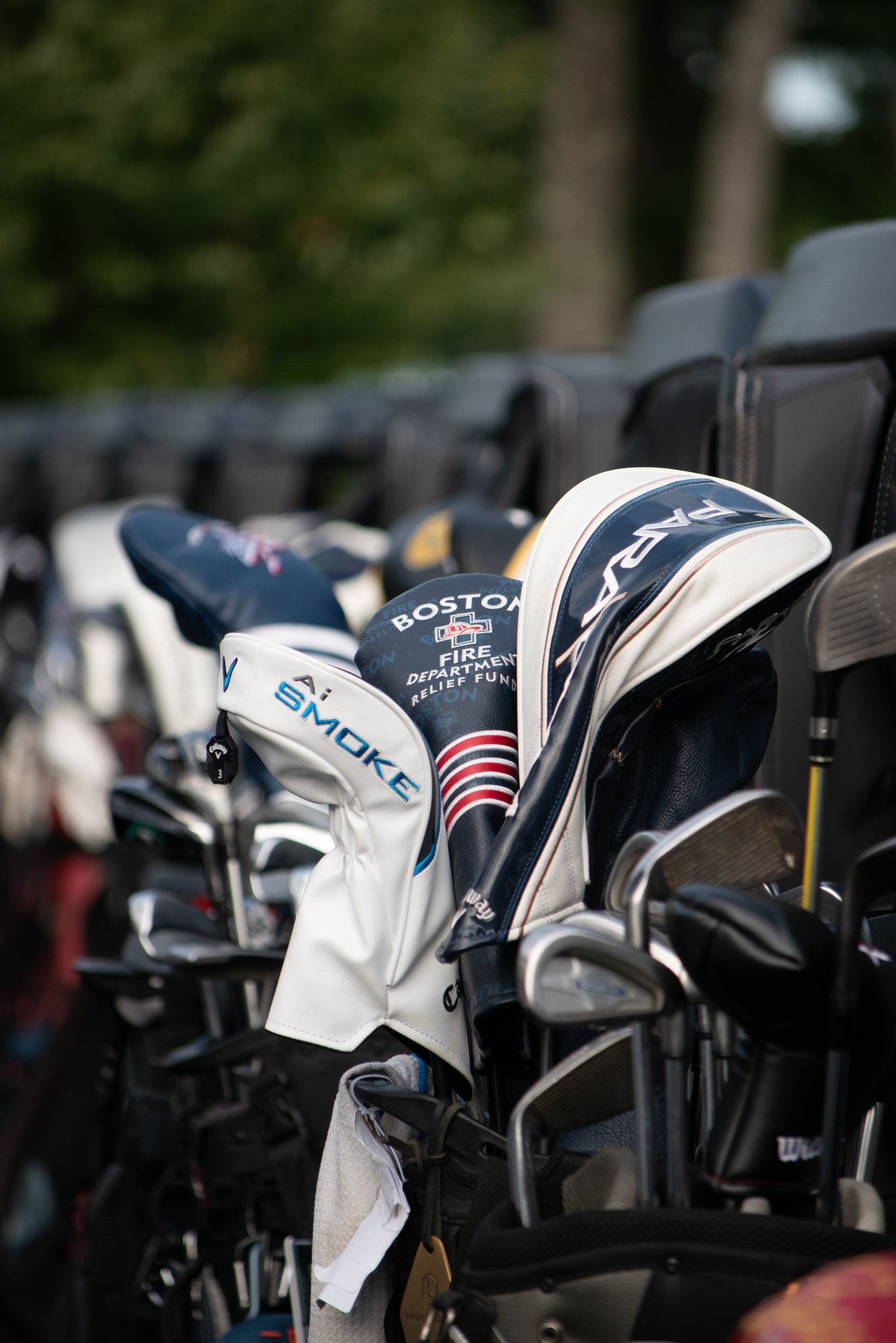 Close-up of golf bags with driver headcovers, including a navy Boston Fire Department Relief Fund cover and a white Callaway Ai Smoke cover.