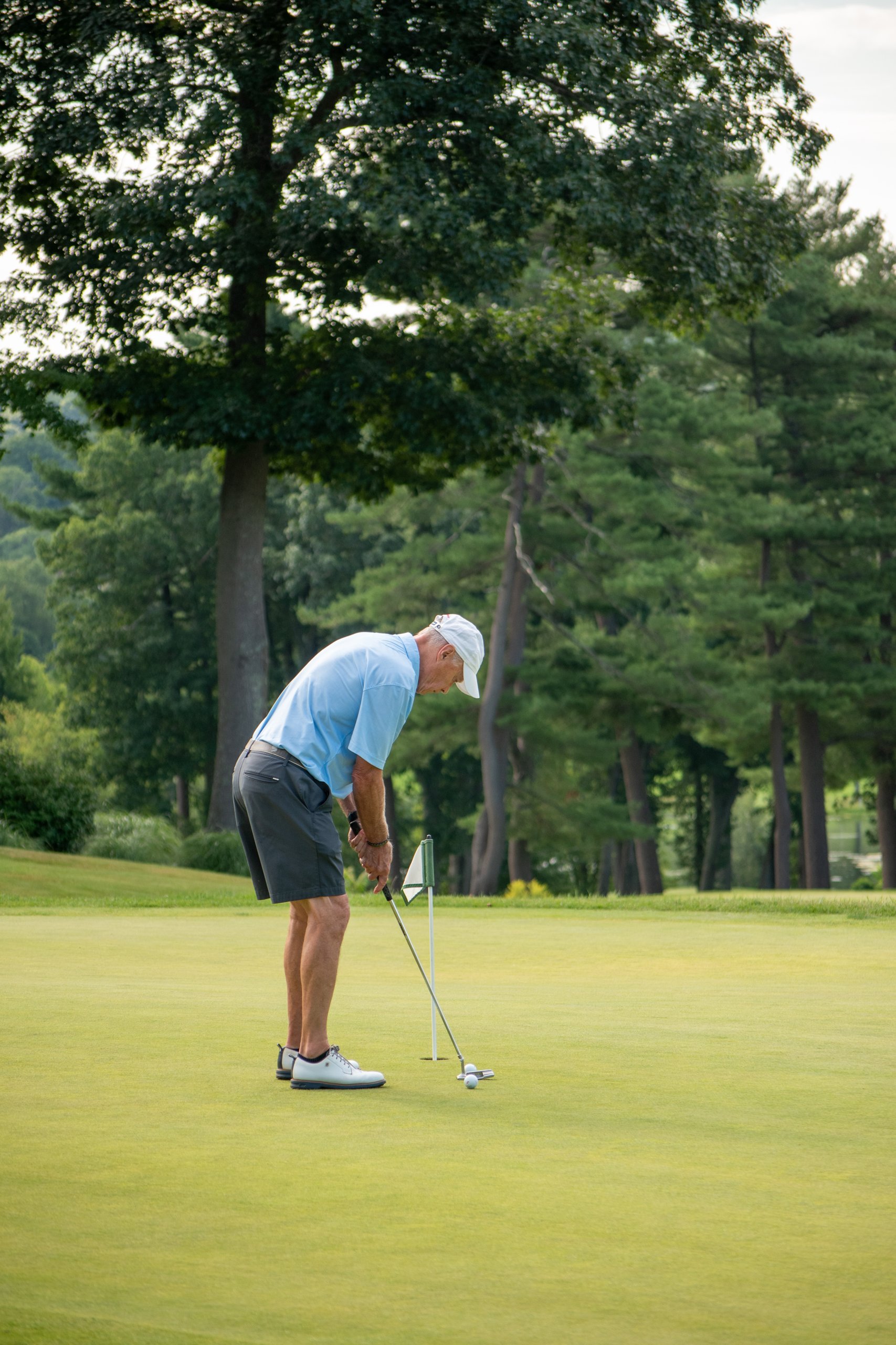 Older golfer in a light blue shirt and white cap putting on the green beside the flagstick, tall trees behind.