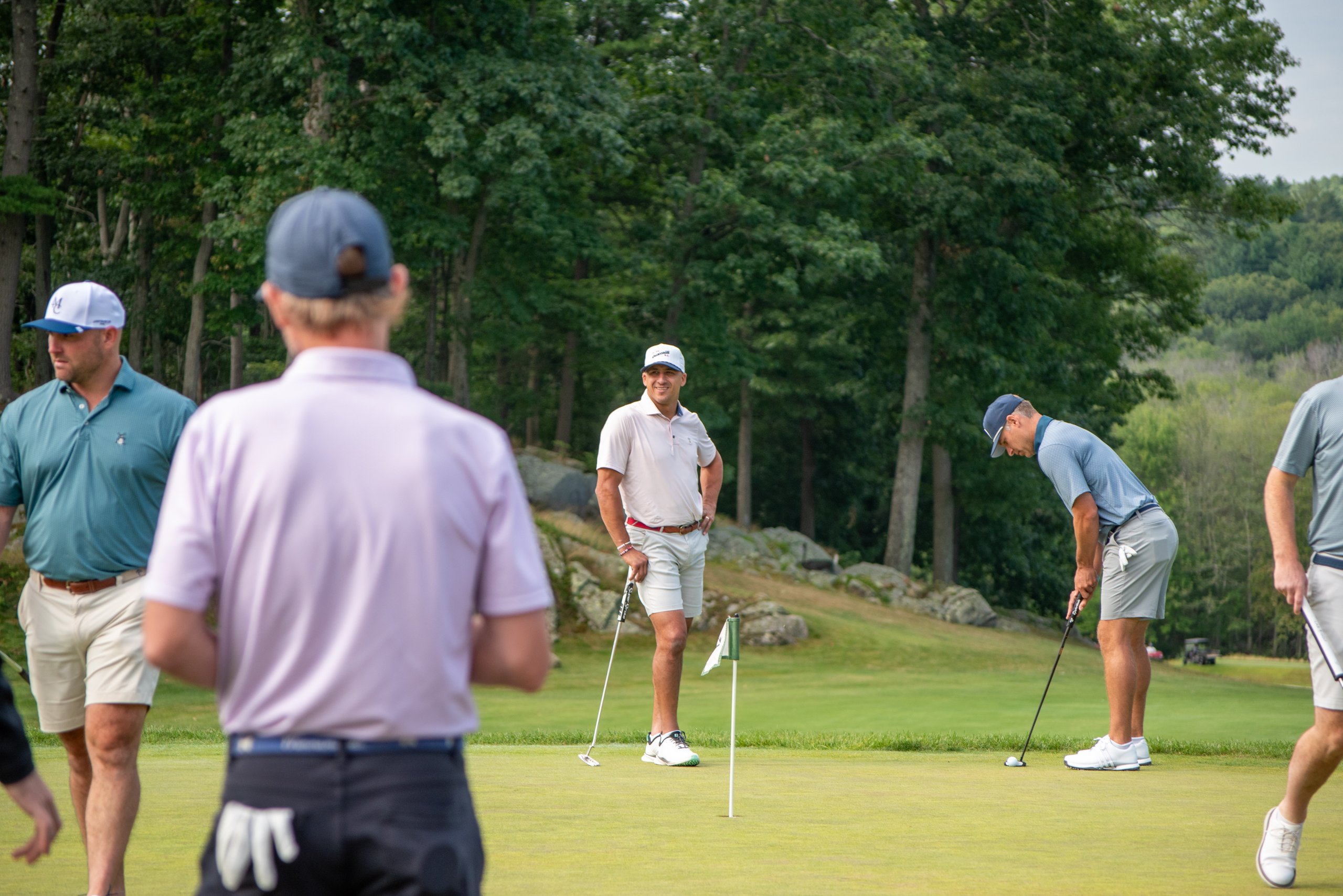 Group of golfers on the green, one bending to putt near the flag while the others watch.