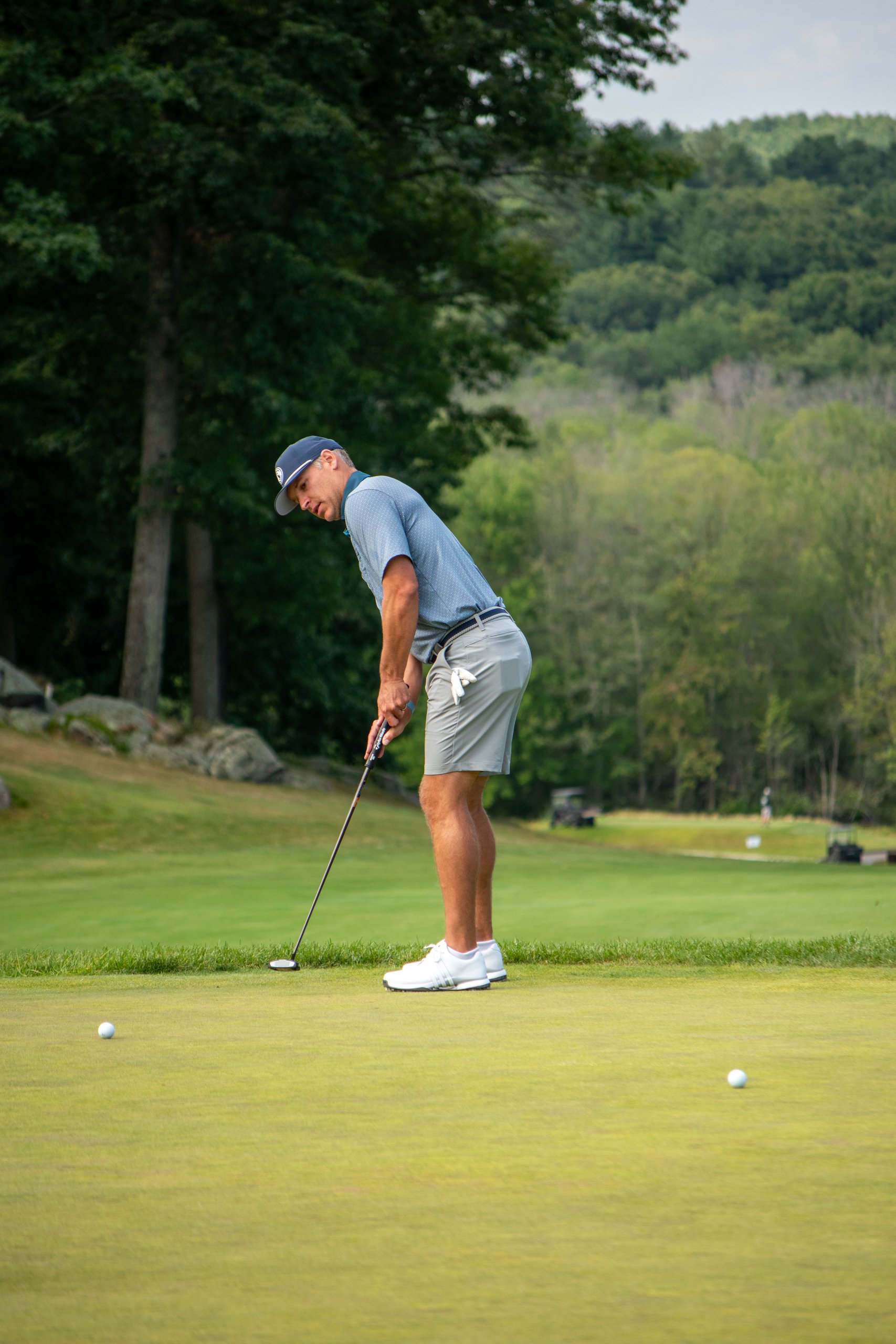 Golfer in a blue polo and navy cap putting on the green, wooded hills in the background.