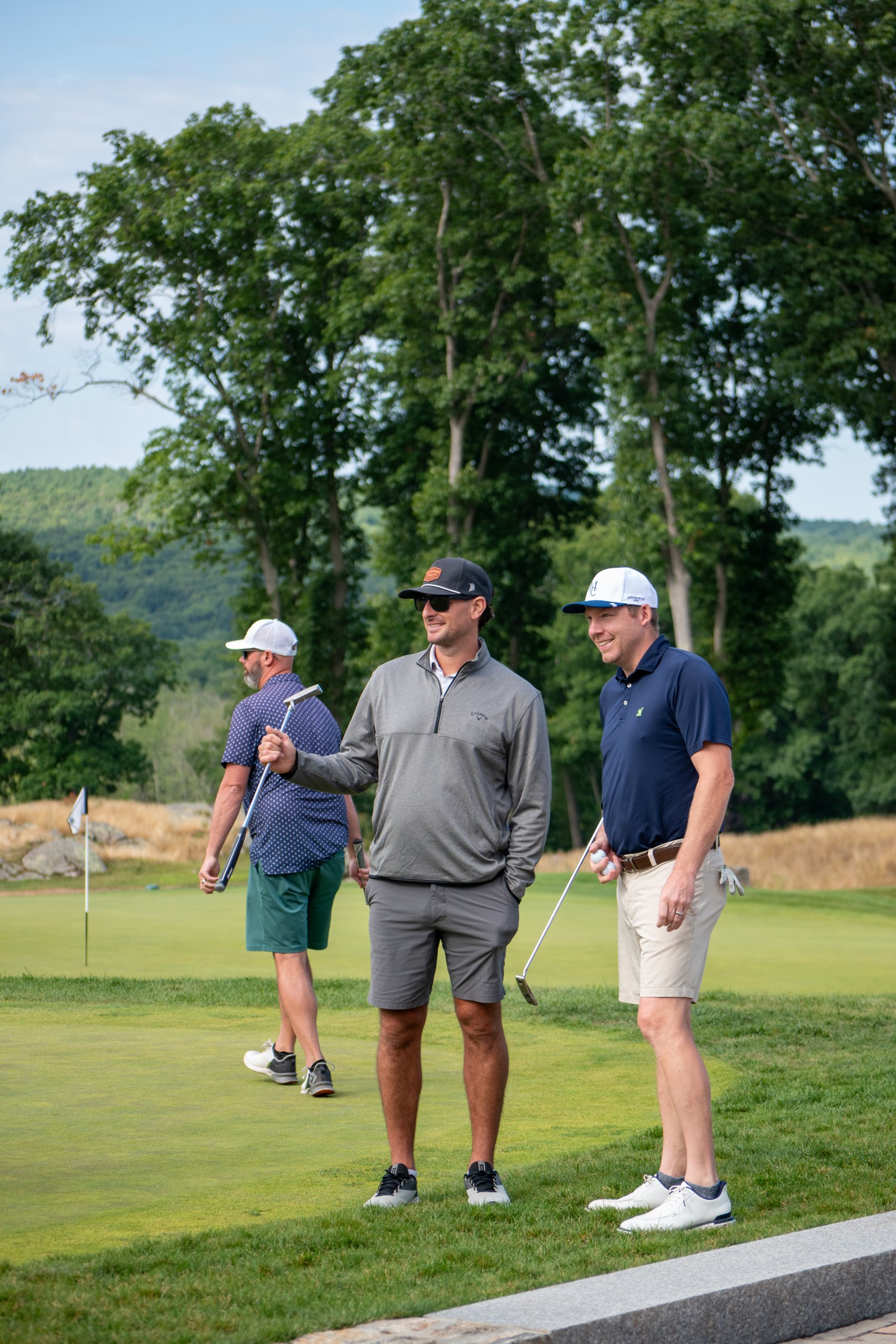 Three golfers standing and talking on the green, one holding a putter, green hills behind.