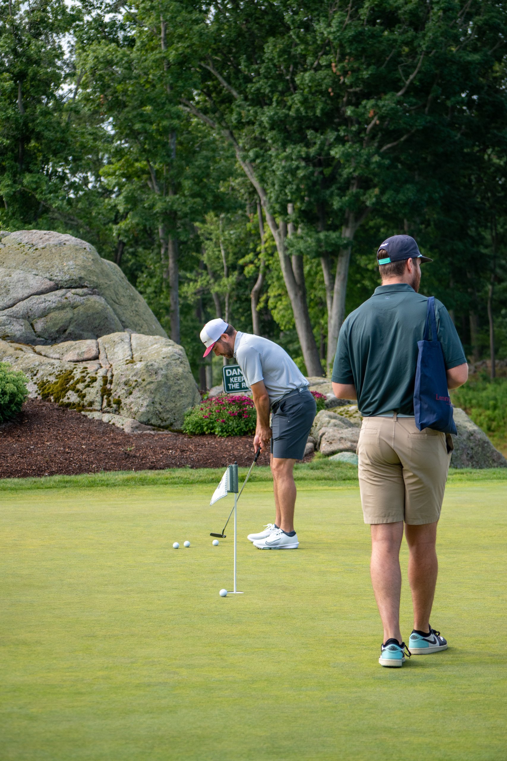 Two golfers on the green lining up putts with several balls near the hole, a large mossy boulder and a navy LennyMade tote behind.