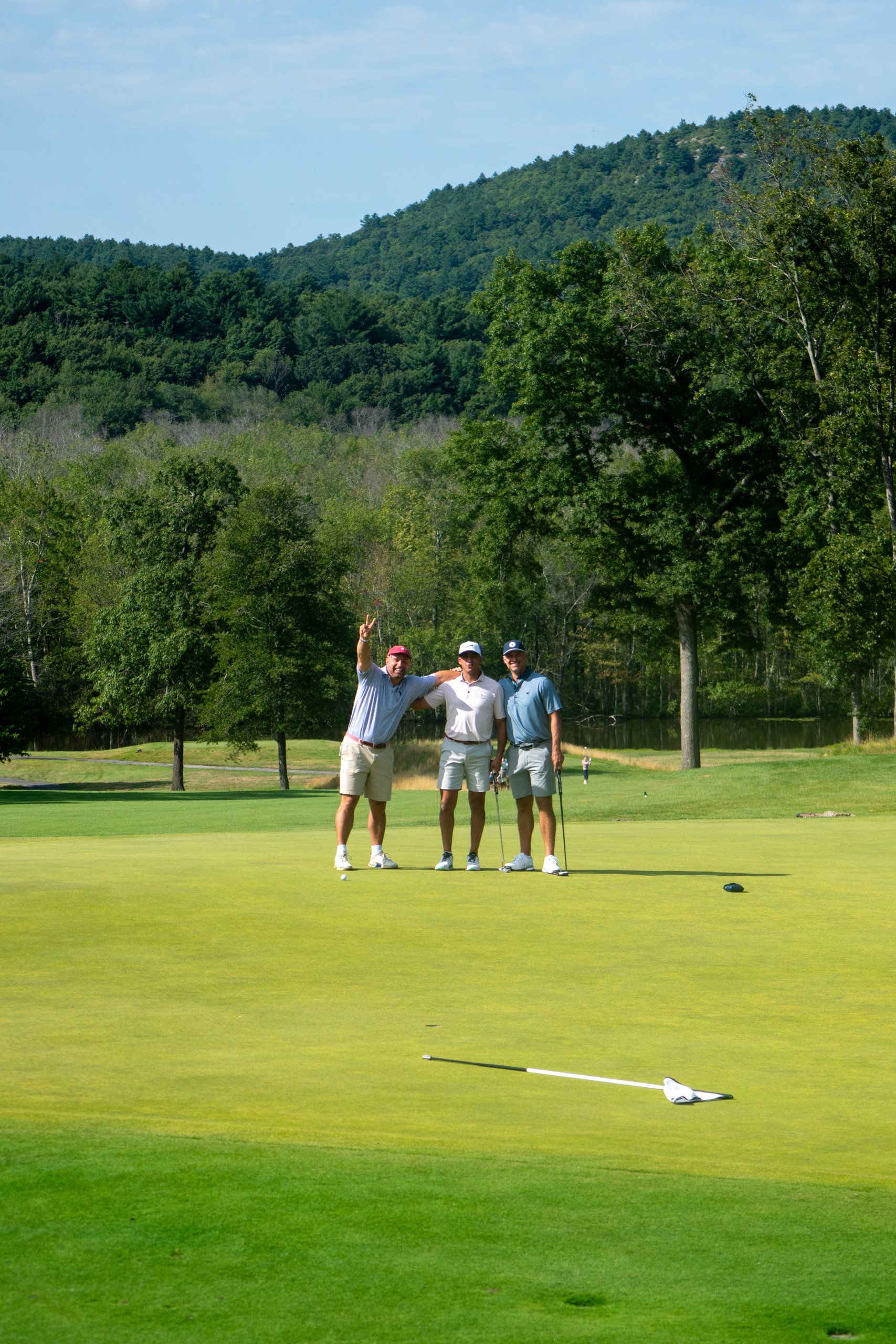 Three golfers posing together on the green, one flashing a peace sign, a wooded hill and pond behind and the flag laid on the grass.