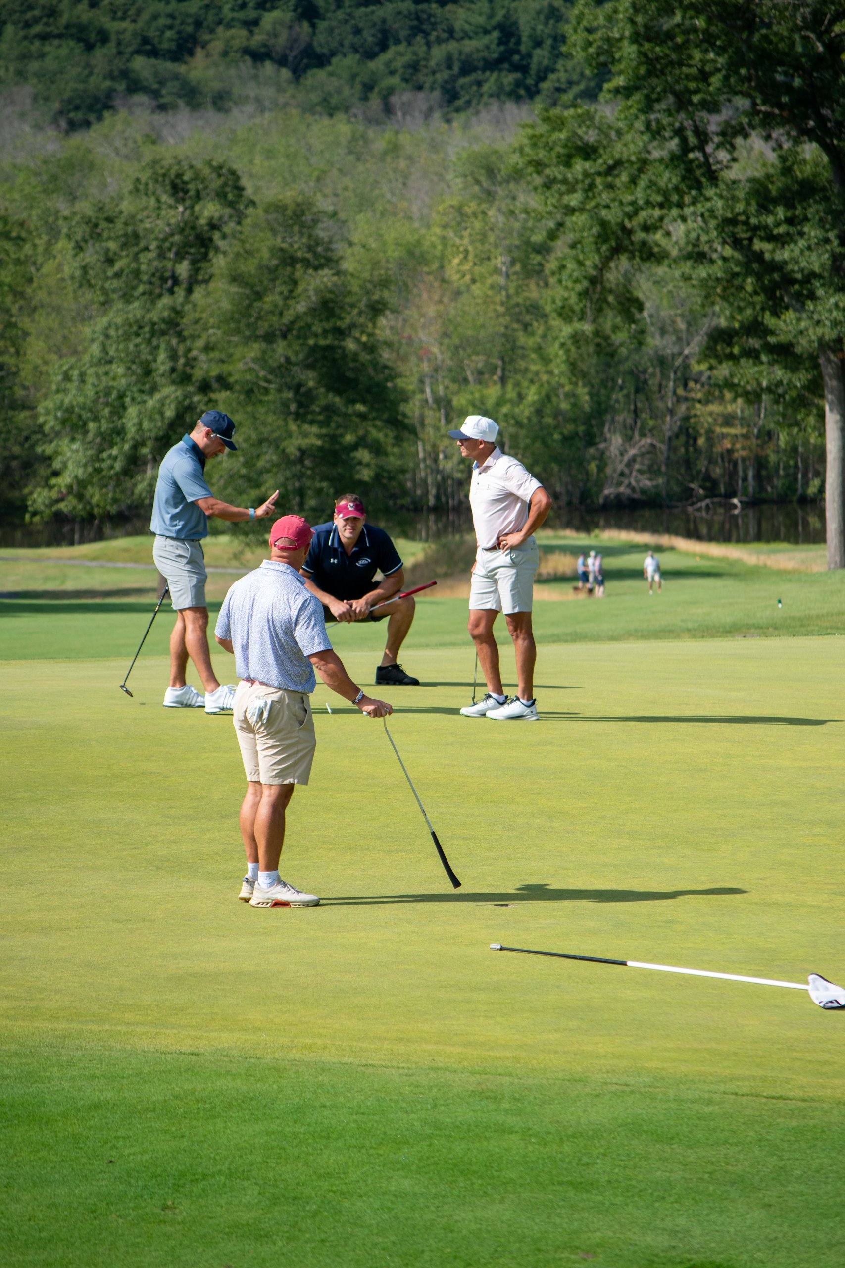 Foursome gathered on the green reading a putt, one player crouching to study the line with the flag laid down.