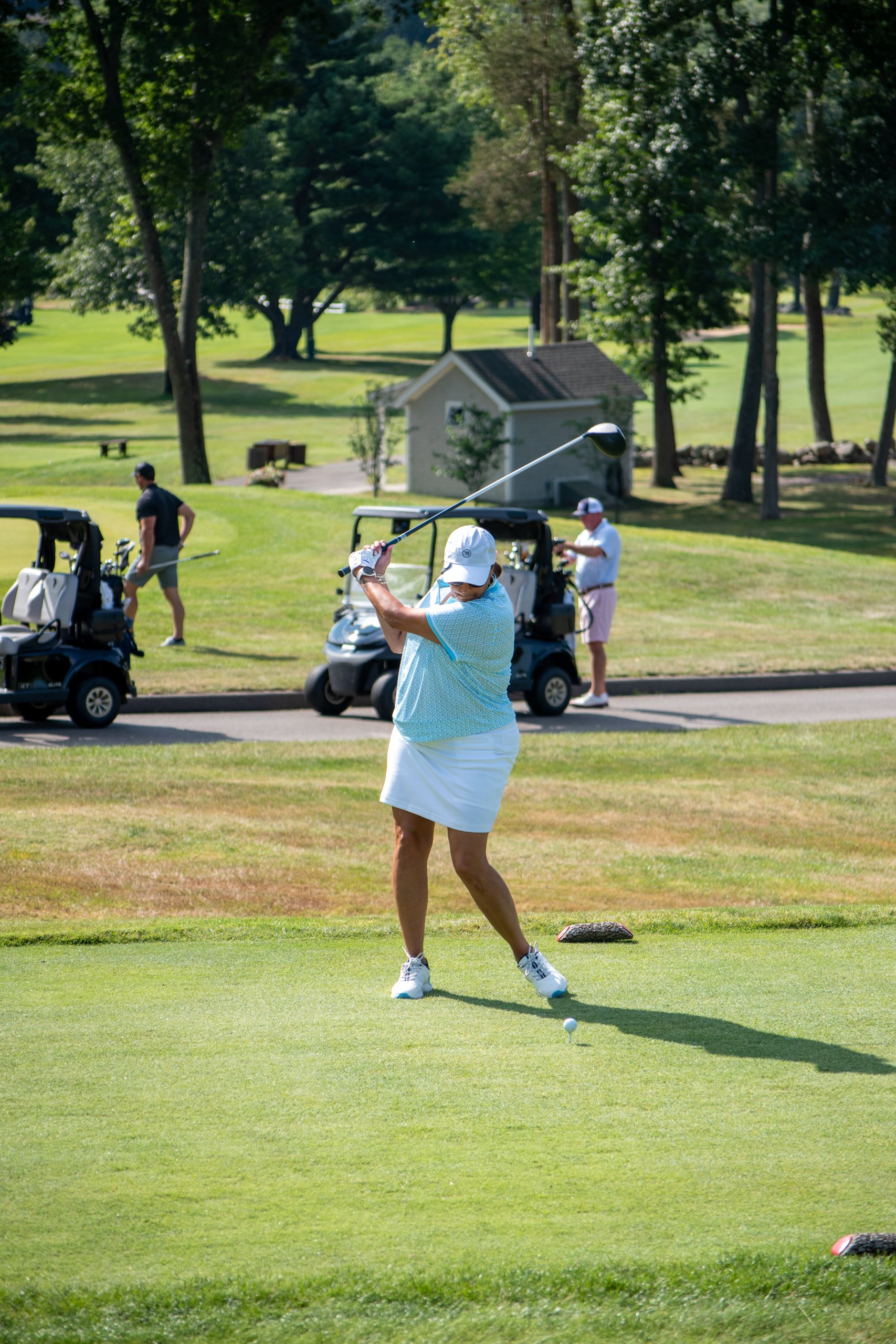 Woman in a teal polo and white skort mid-swing driving off the tee, golf carts and a course shed behind.