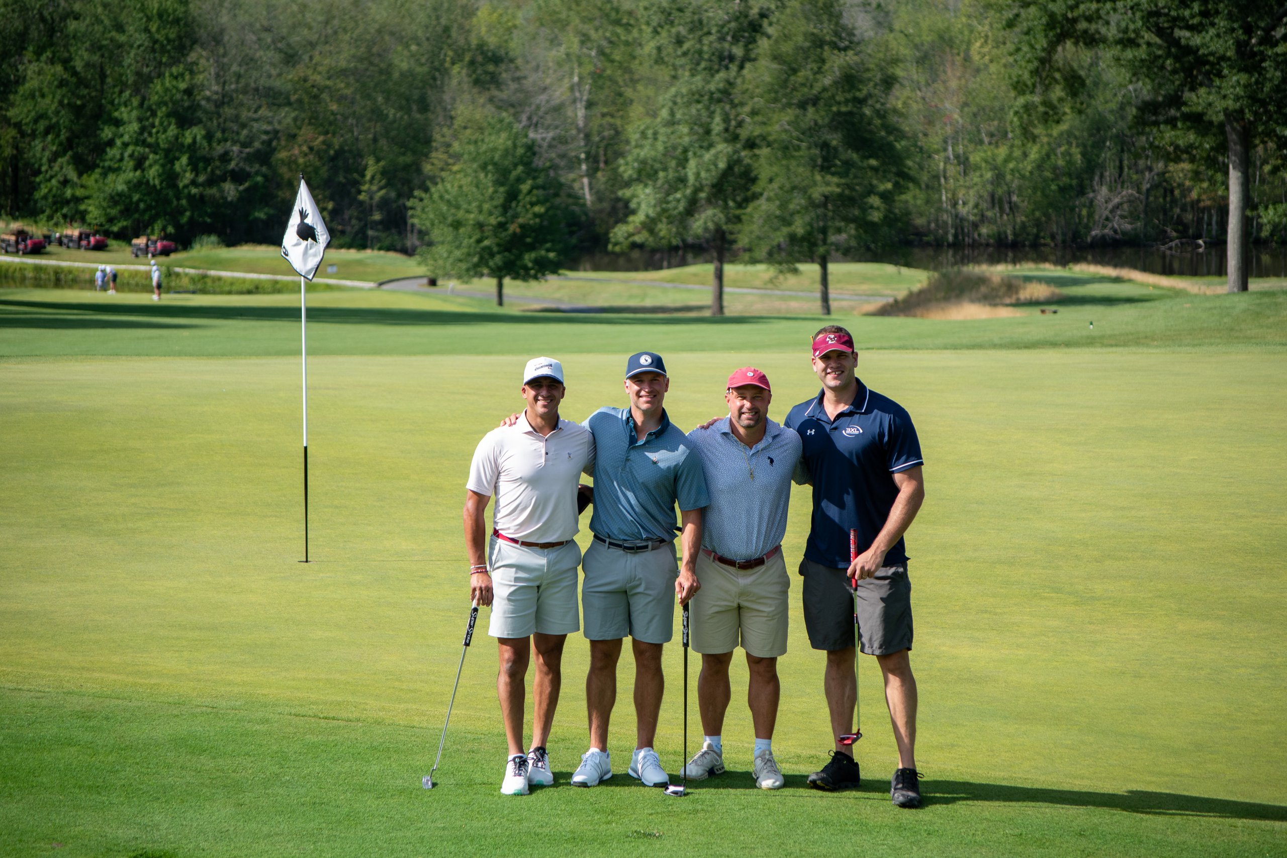 Foursome of golfers posing together on the green beside the flag, a pond and wooded hills behind them.