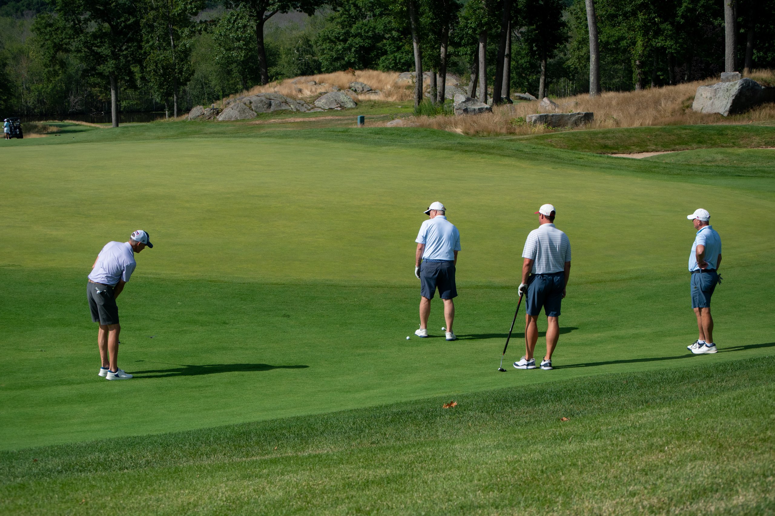 Four golfers spread across the green, one lining up a putt while the others watch, a rocky outcrop and tall grass behind.
