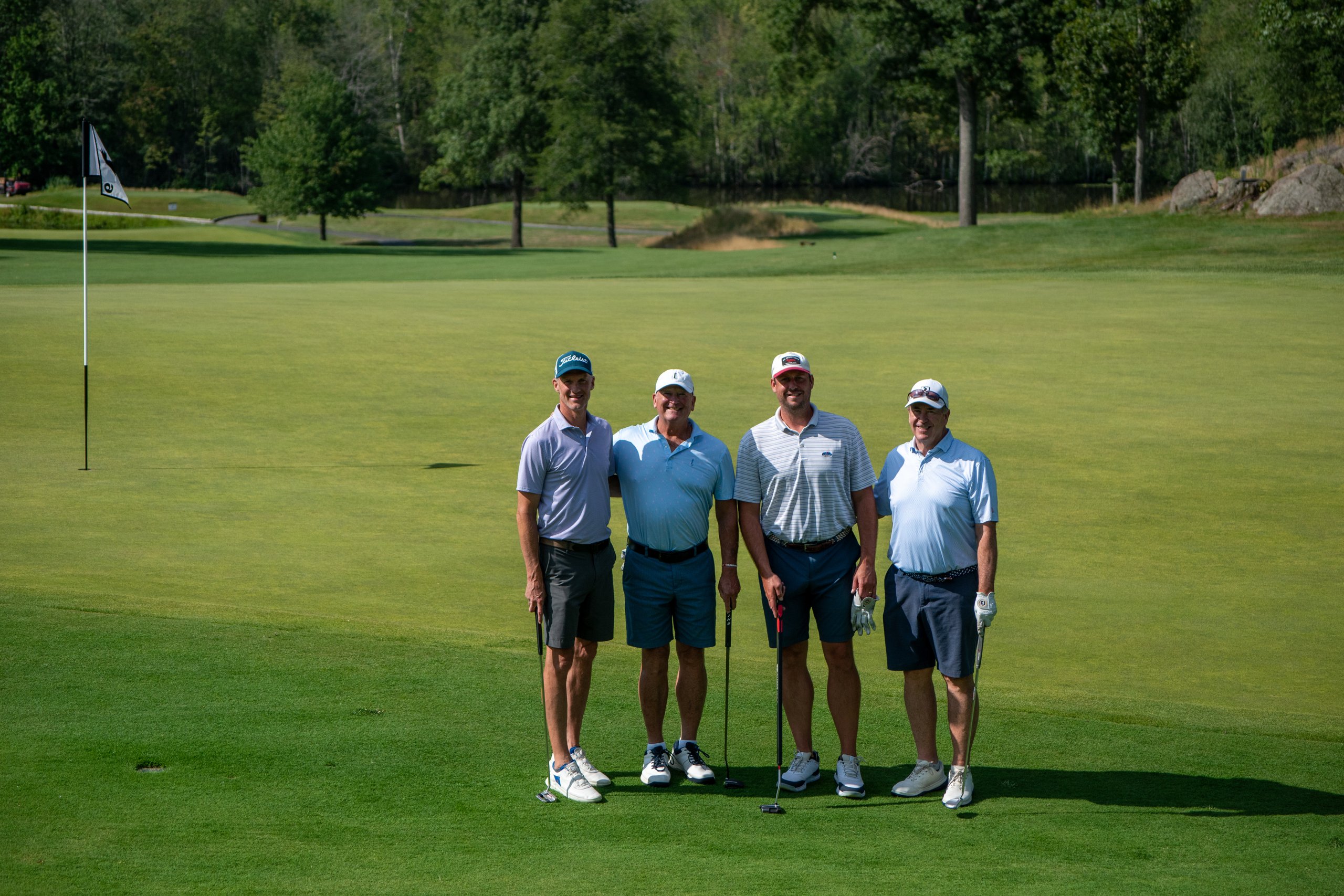 Foursome of golfers posing together with their putters on the green, the flag and fairway behind them.