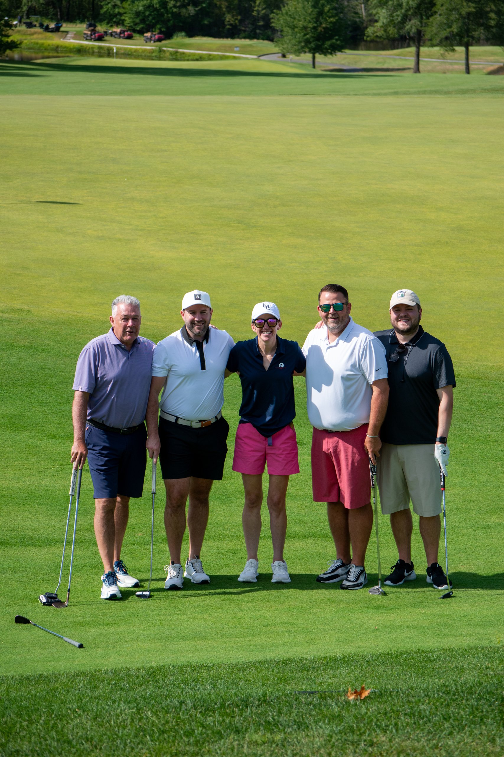 Same group of five golfers posing together on the green, golf carts and a pond in the distance behind them.