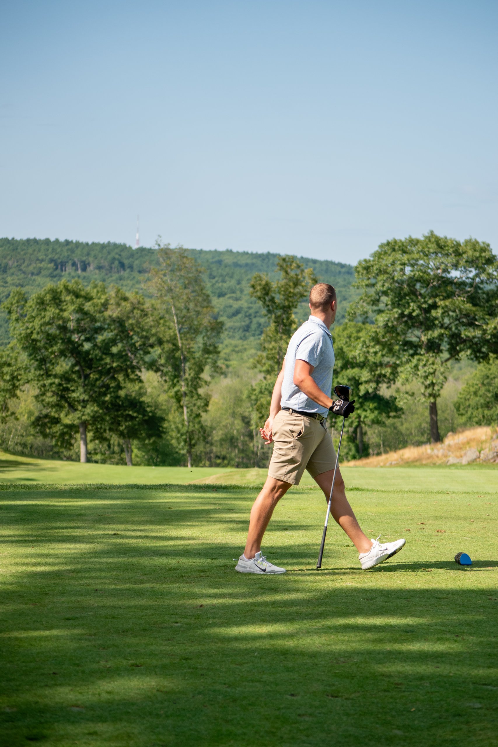 Golfer in a light blue shirt and khaki shorts walking across the tee carrying a driver, a forested hill behind.