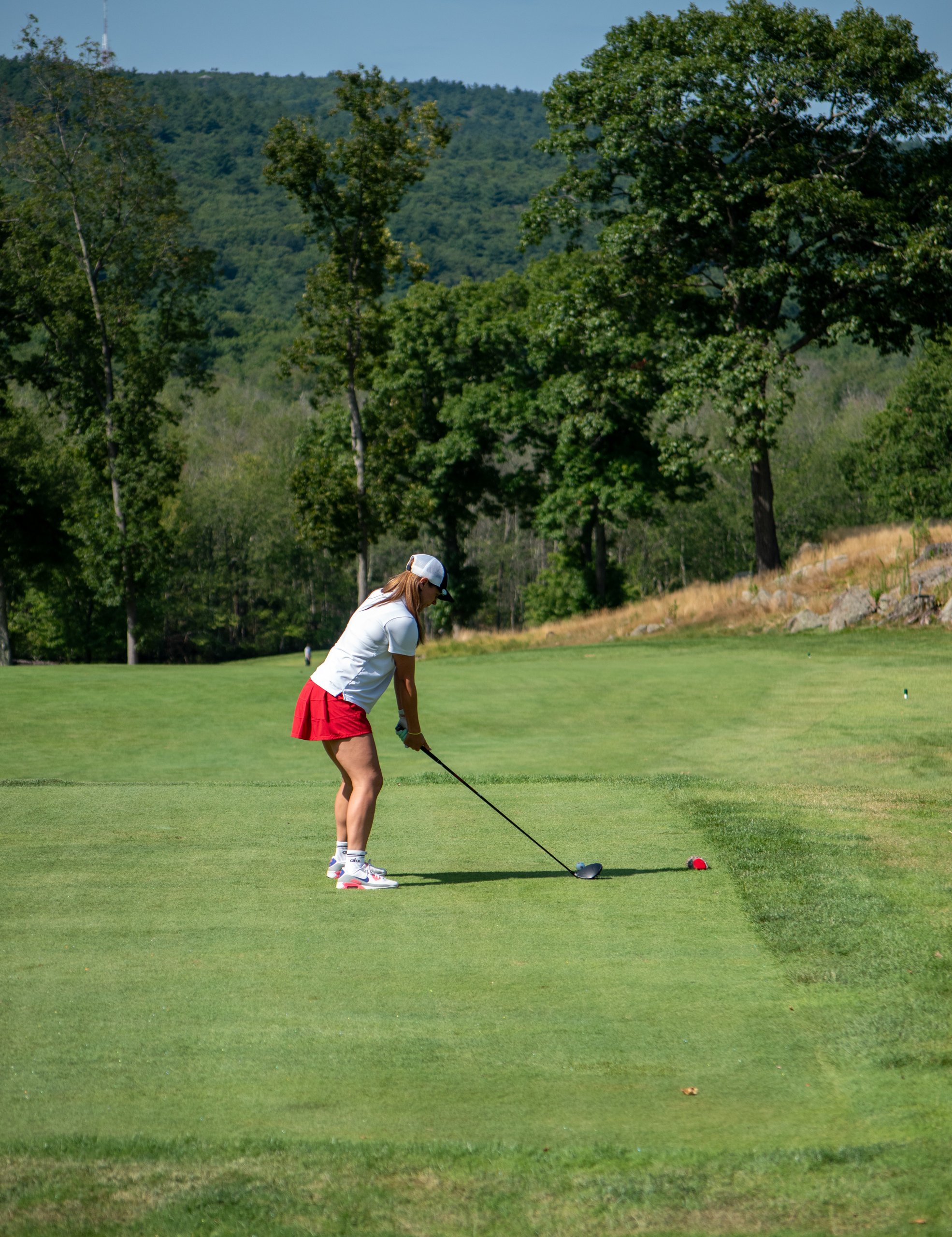 Woman in a white top and red skort teeing off with a driver, a wooded hillside behind the tee.