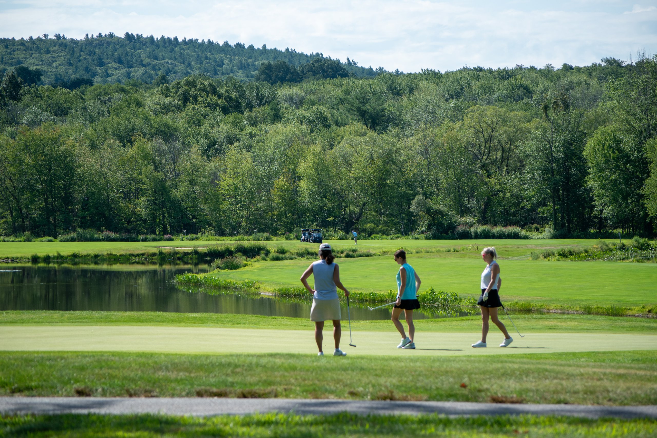 Three women walking across the green with their putters beside a pond, a forested hill in the background.