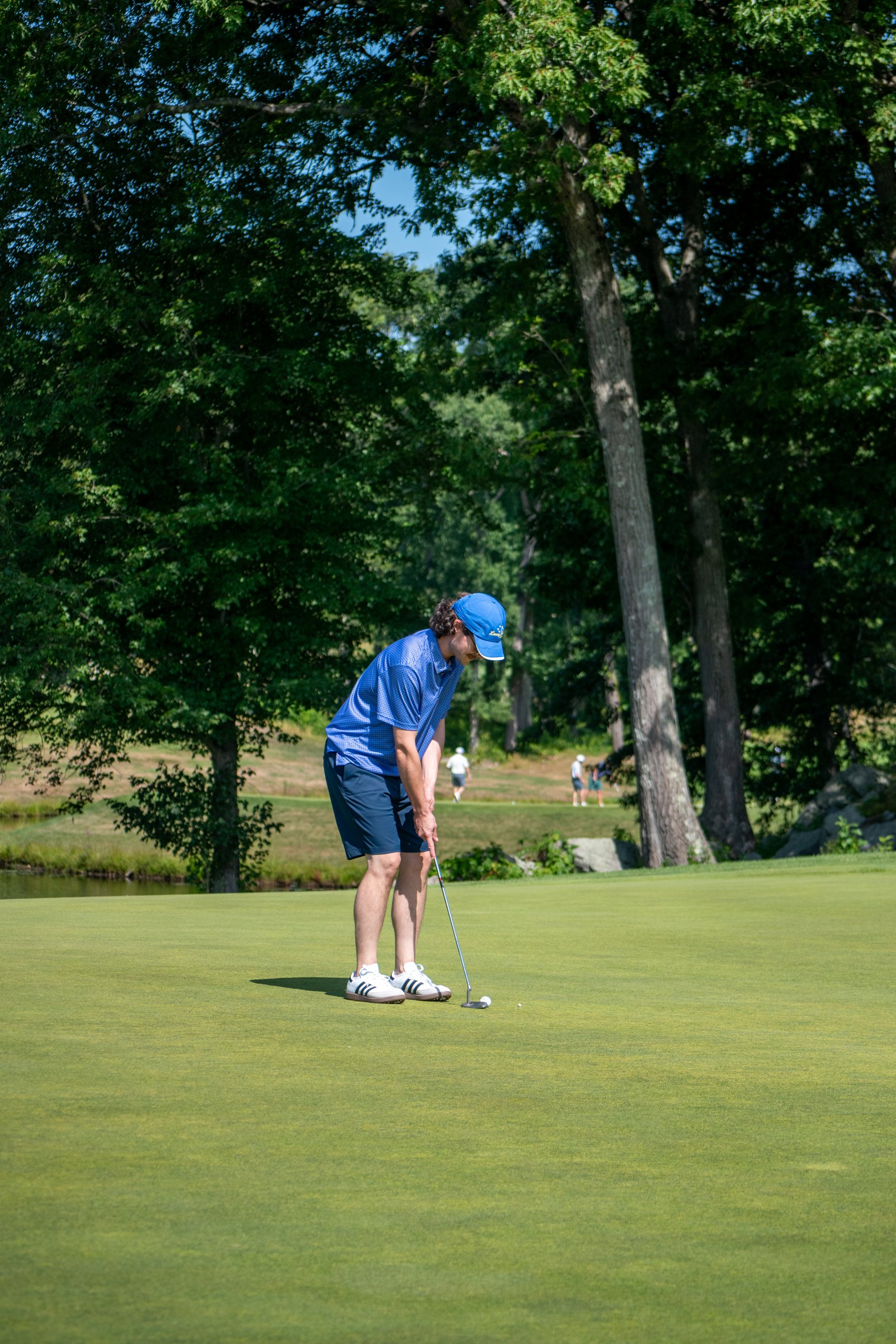 Golfer in a blue polo and blue cap putting on the green, trees and a pond behind.