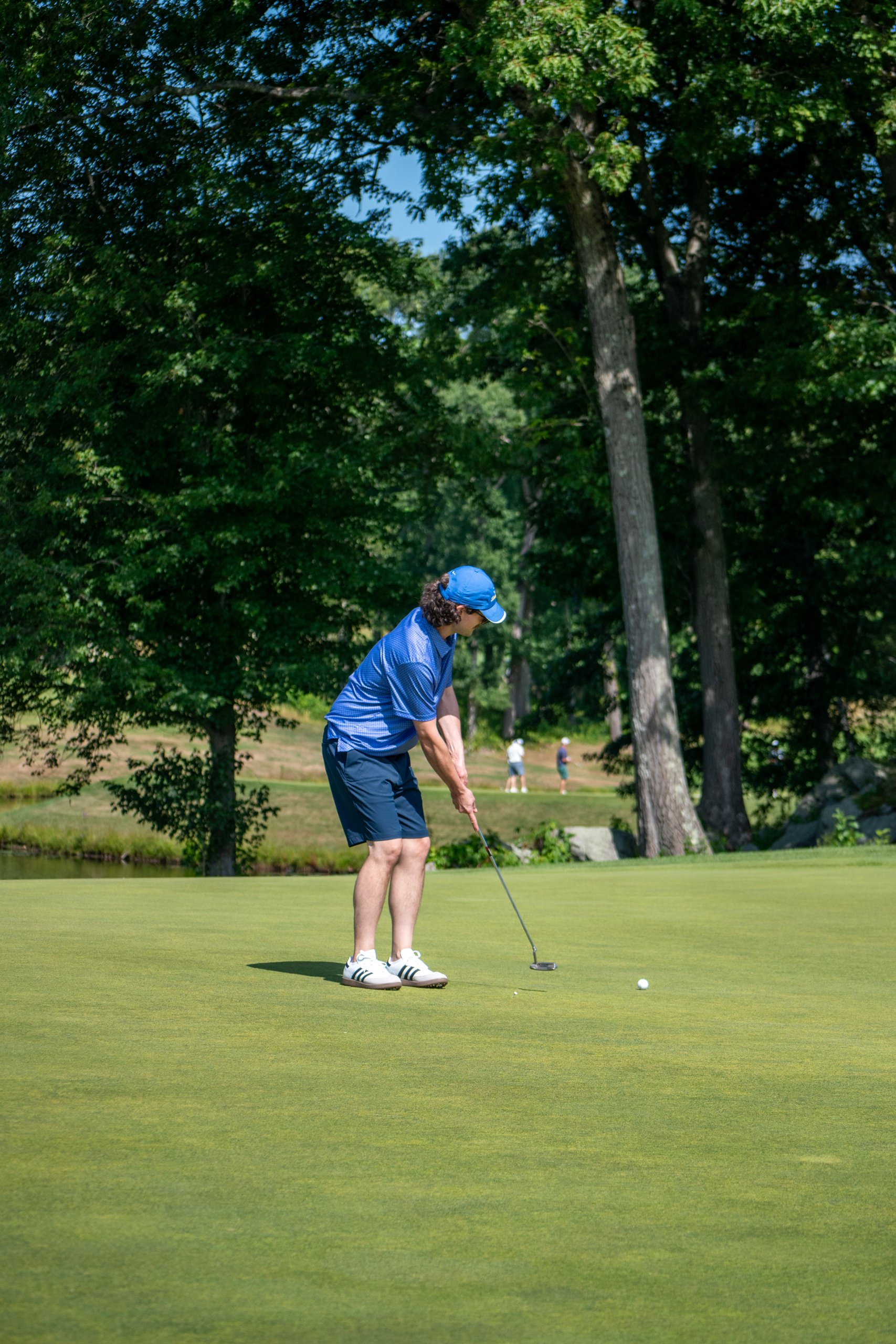 Golfer in a blue polo and cap stroking a putt on the green, other players and a pond in the background.