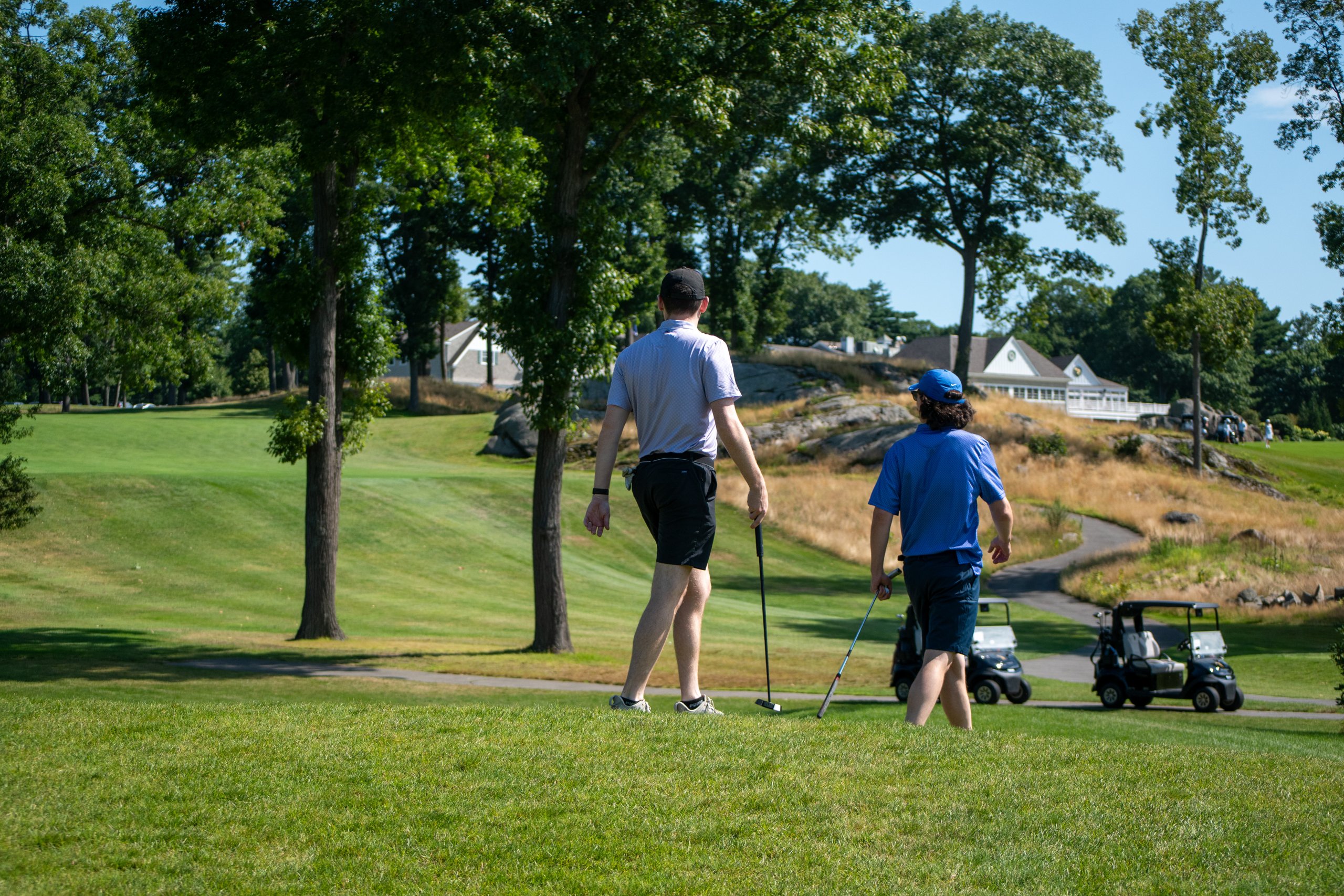 Two golfers walking up the fairway with their putters toward the clubhouse on the hill, golf carts on the path beside them.