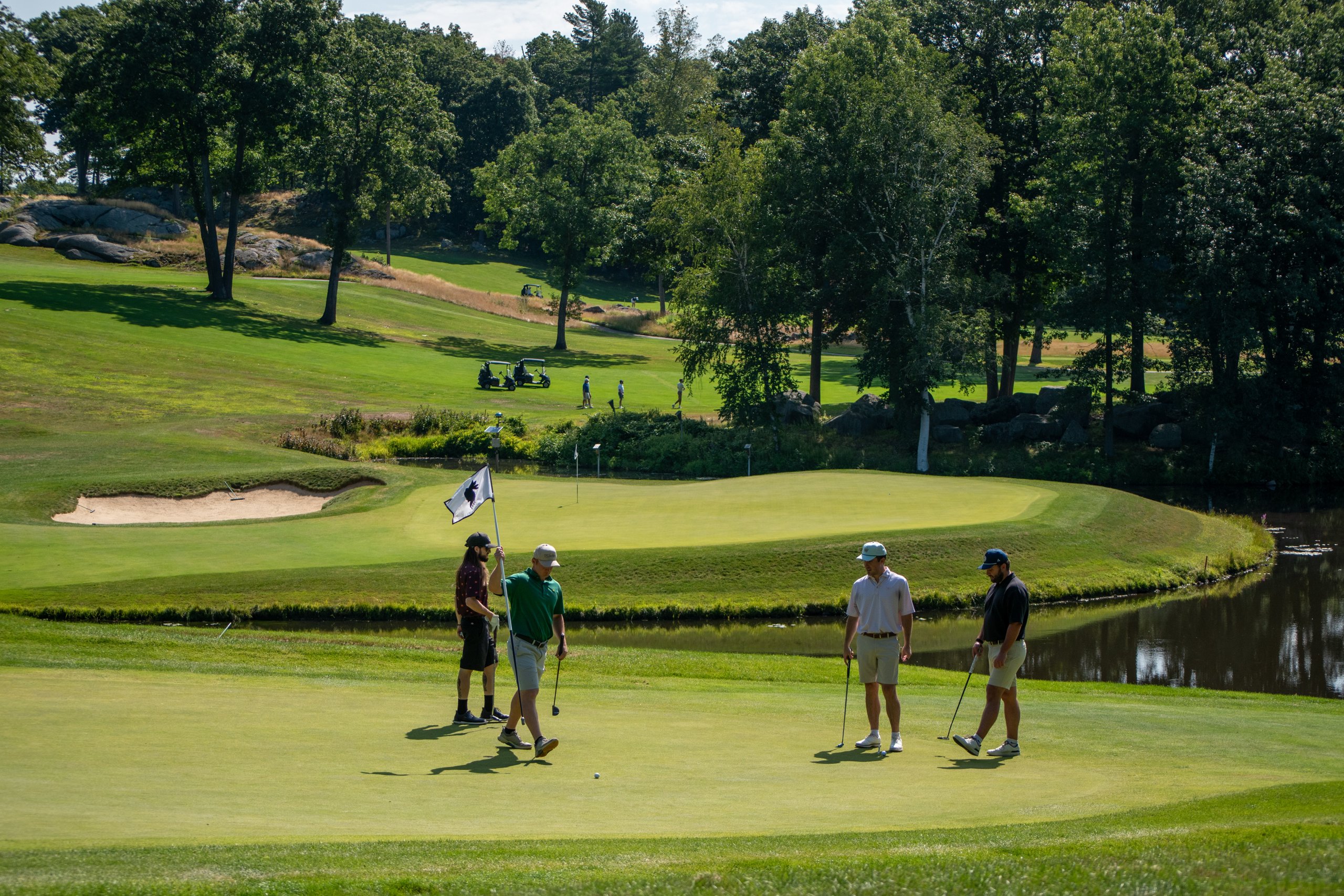 Four golfers on the green near the pin, one holding the flag, with a pond and rocky wooded hillside behind.