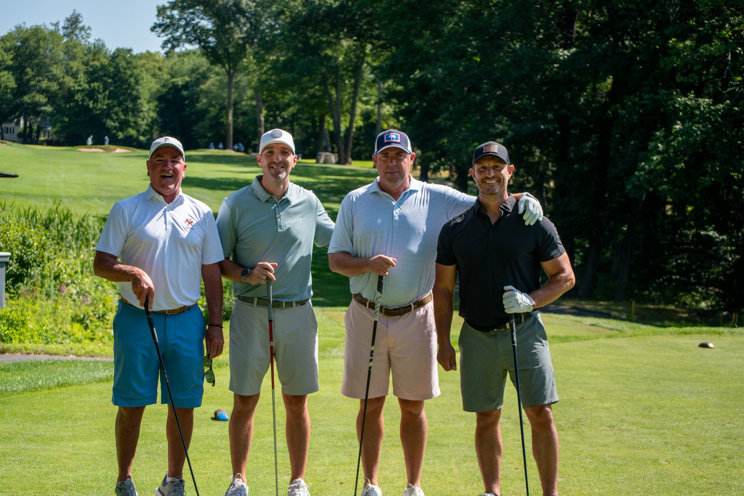 Four men posing shoulder to shoulder on the tee with their clubs, smiling for the camera.