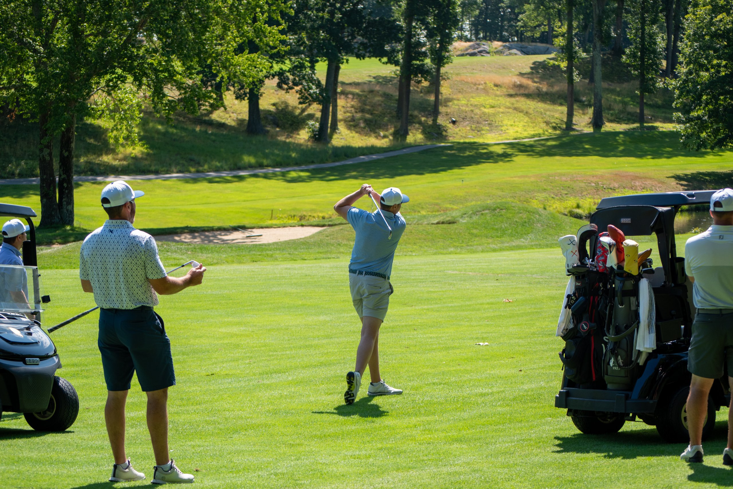 Golfer in a blue polo following through on an iron shot from the fairway as a teammate watches beside the cart.