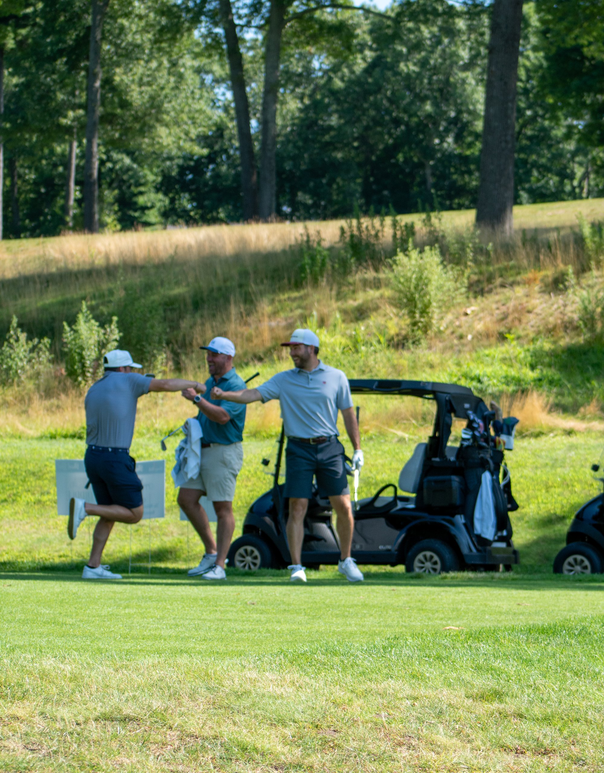 Three golfers celebrating with a fist bump beside their carts on the fairway, a tall grassy bank behind.