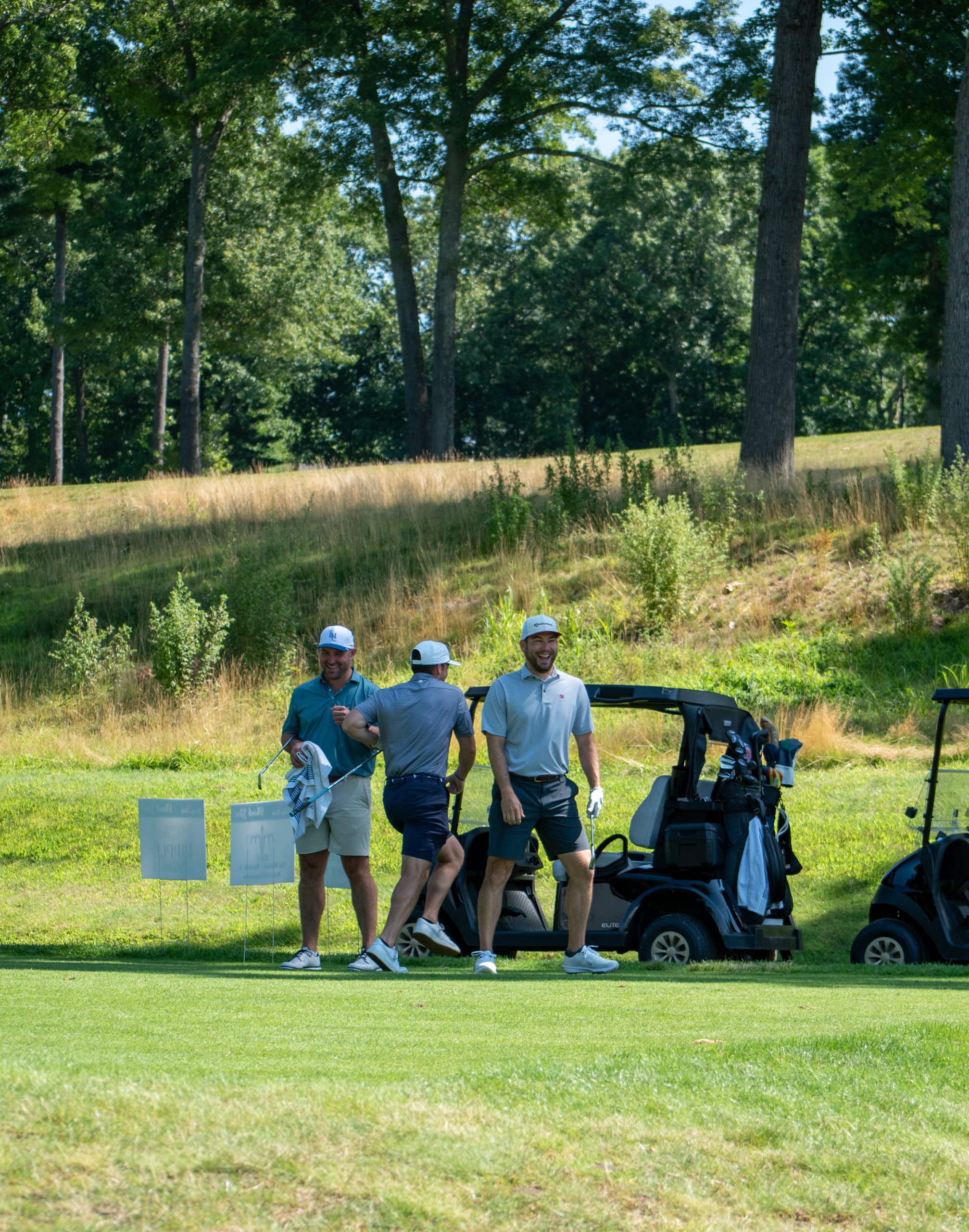 Three golfers laughing and fist-bumping by their carts near sponsor tee signs, a grassy hill behind.
