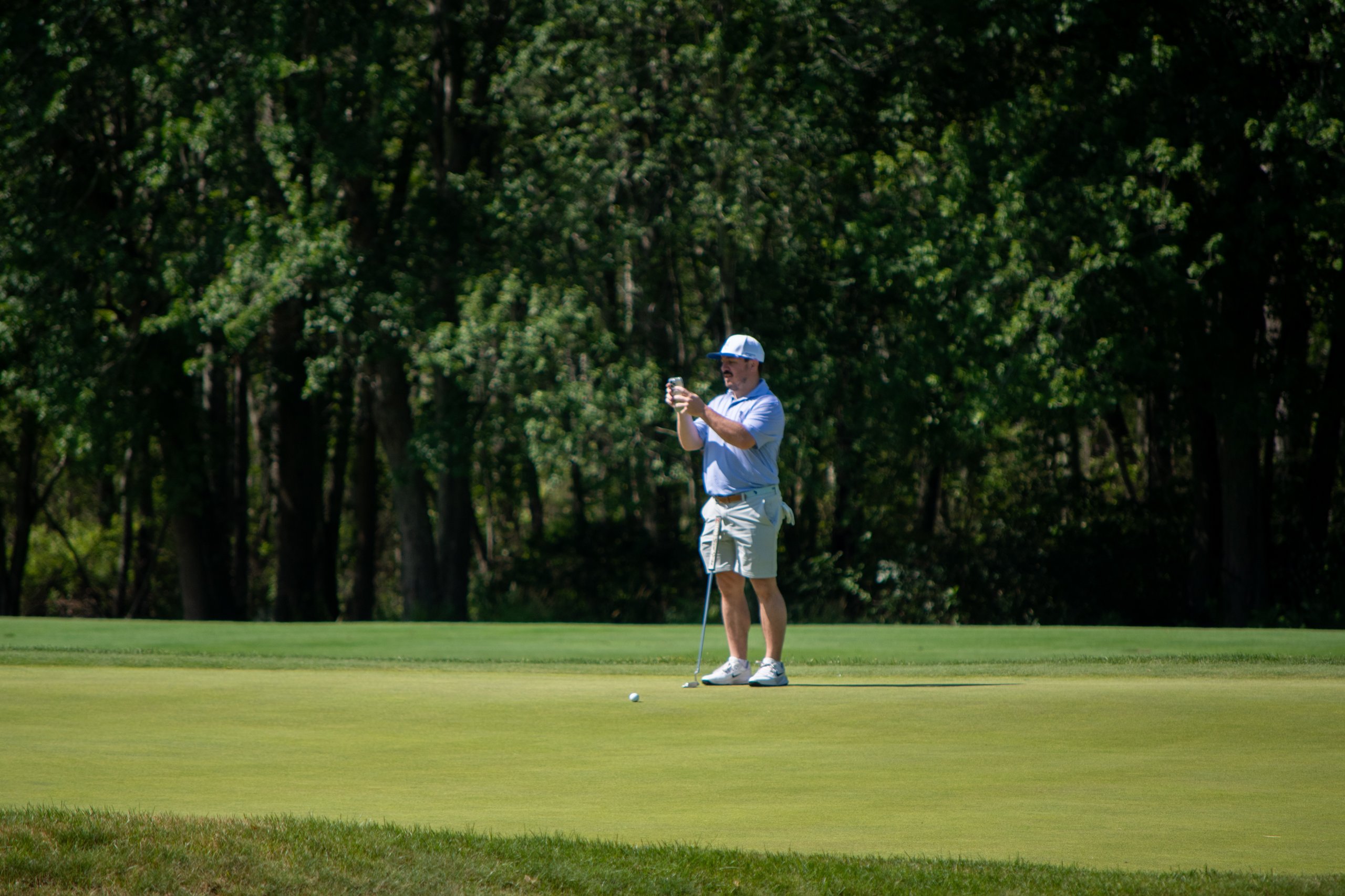 Golfer in a light blue polo standing over his ball on the green holding up a phone, dark trees behind.
