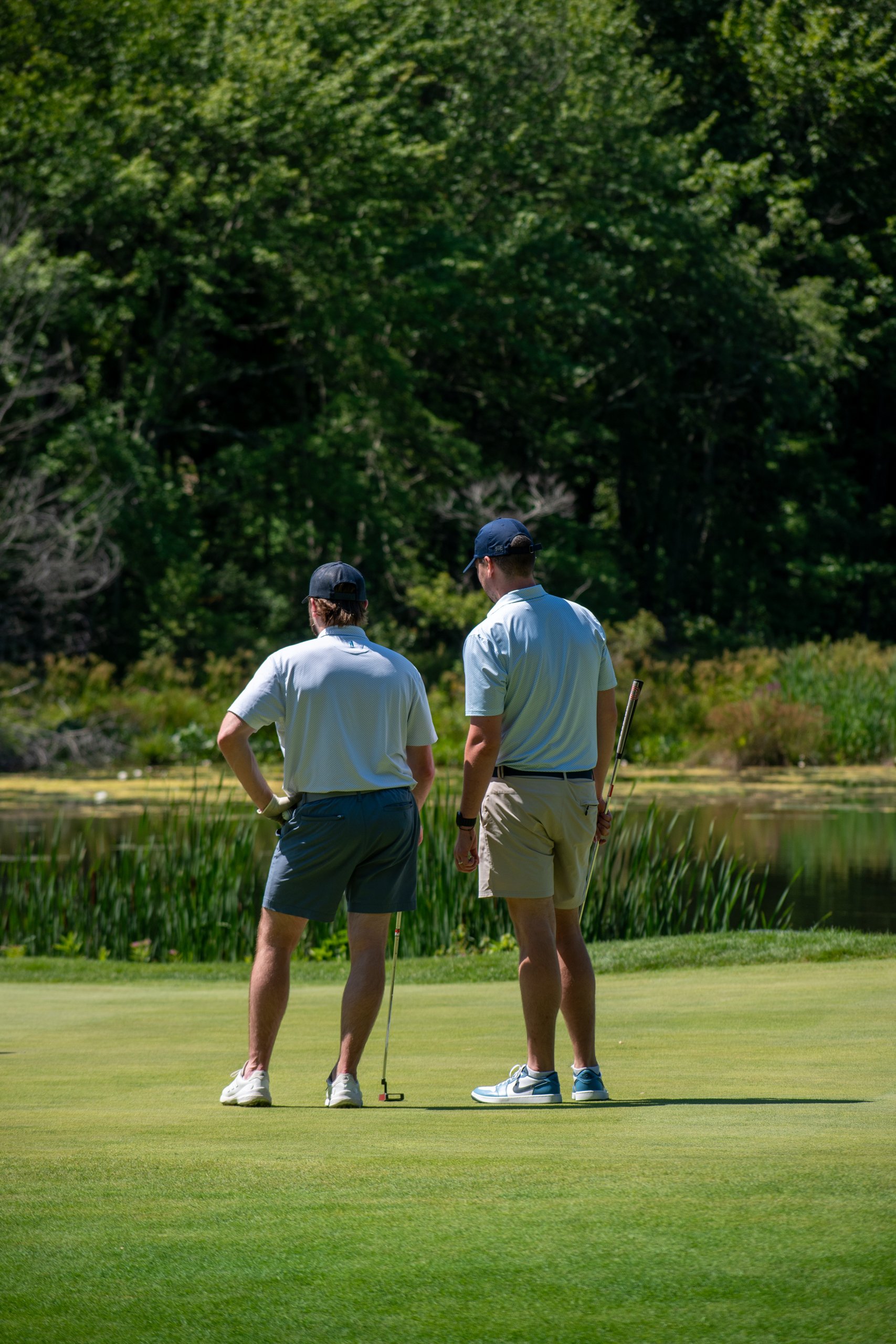 Two golfers standing on the green with their putters, looking out over a cattail-lined pond.