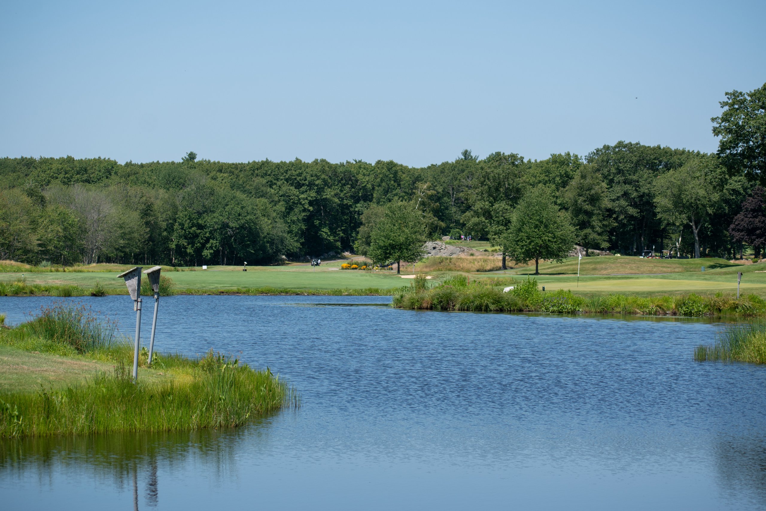 Calm pond with two aerator pipes in the foreground and a green with the flag across the water, forest behind.