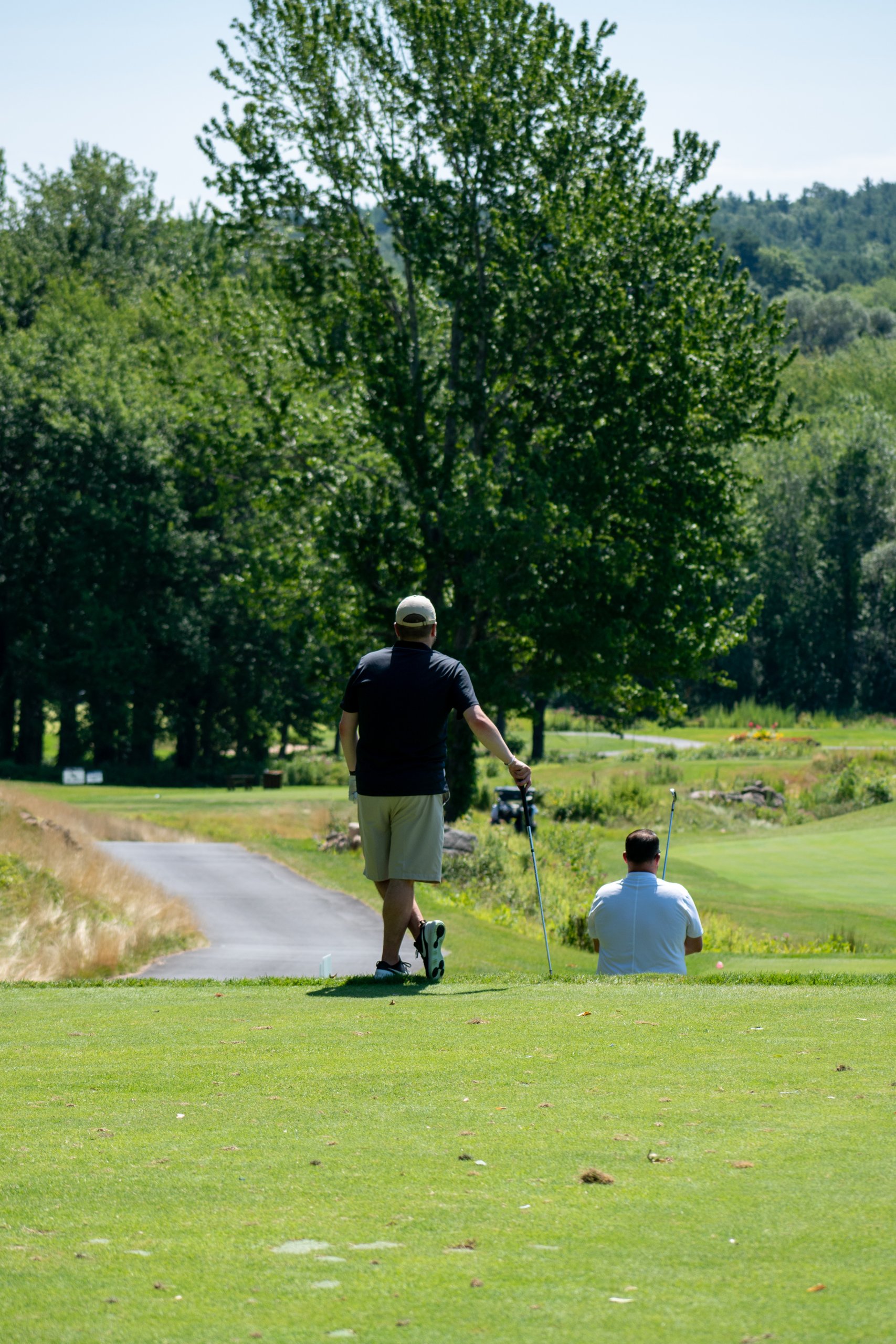 Two golfers walking down a tree-lined tee toward the fairway, one in a black polo carrying a club.