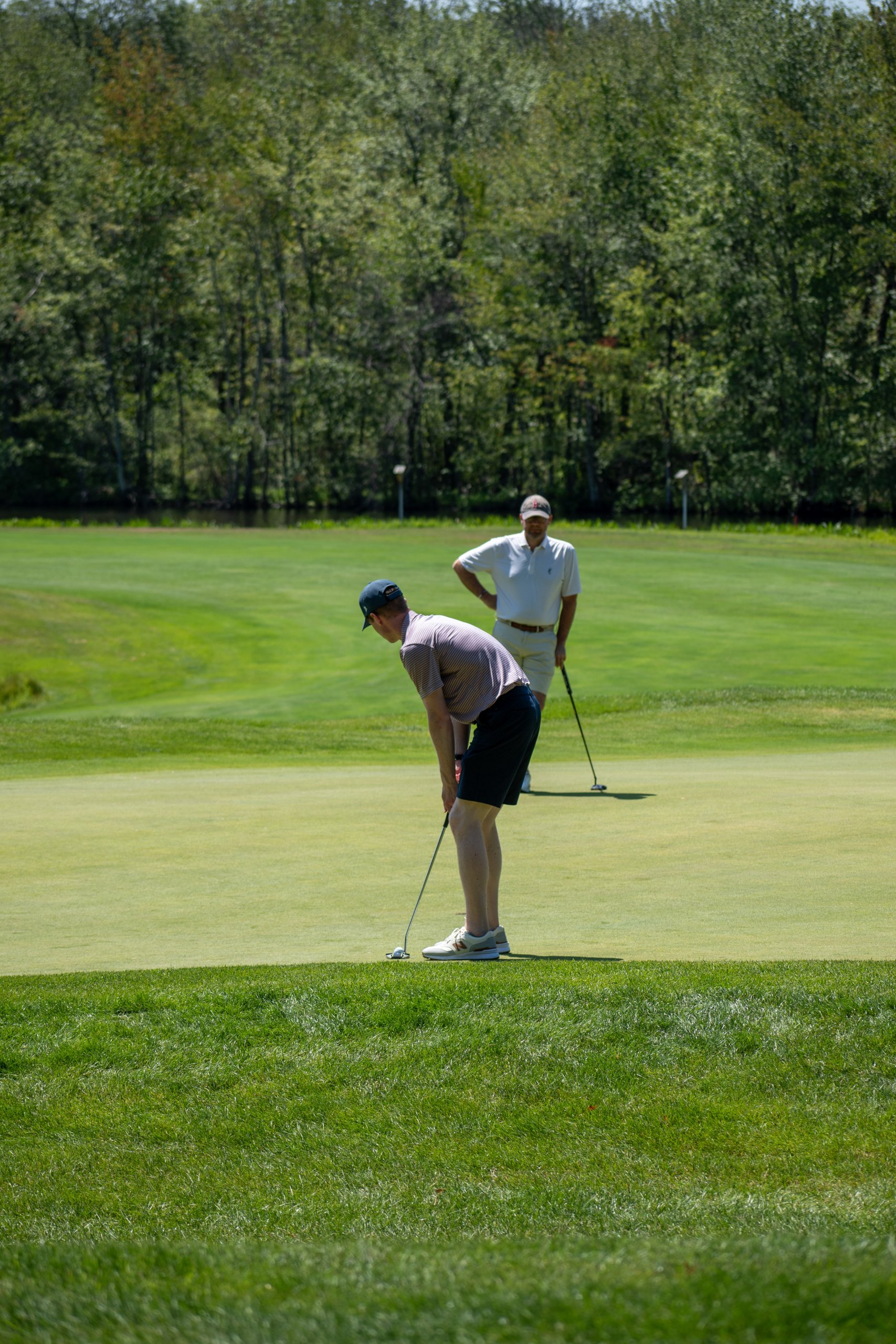 Golfer in a striped polo bent over a putt on the green while a teammate in white watches from behind.