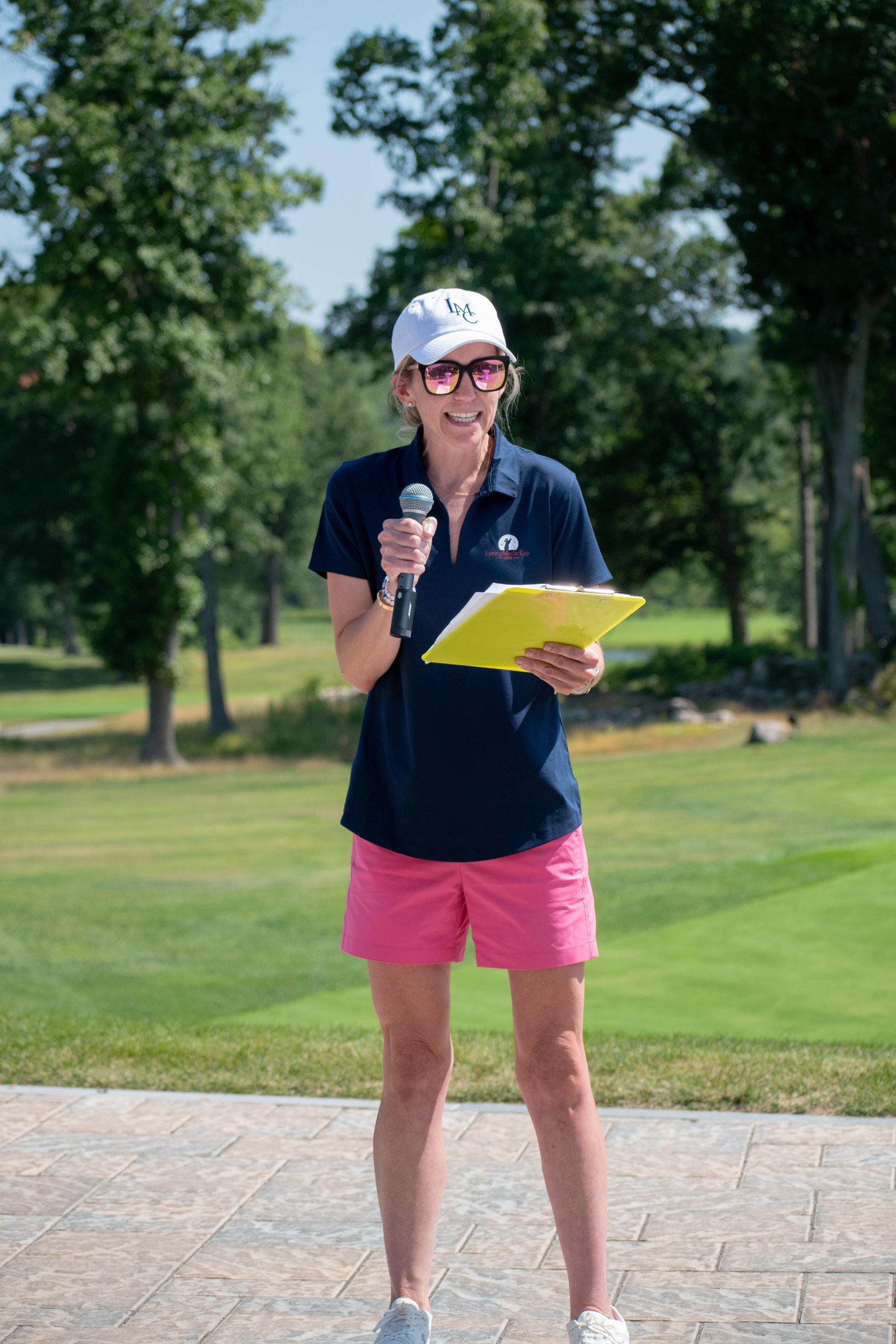 Event host in a LennyMade Cup polo and white LMC cap speaking into a microphone while holding a yellow clipboard on the course.