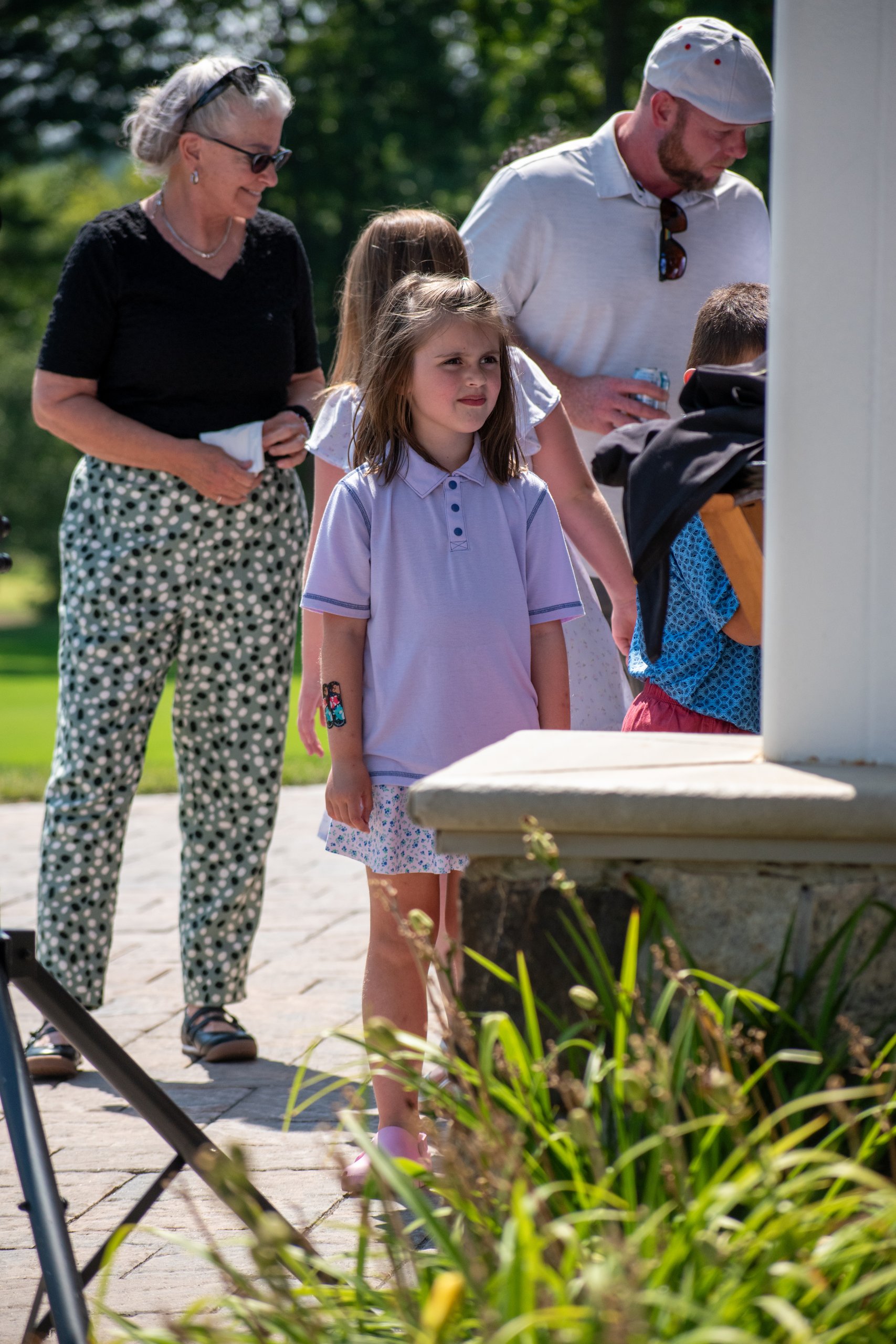 A young girl in a lavender polo standing on the clubhouse patio with family members behind her.