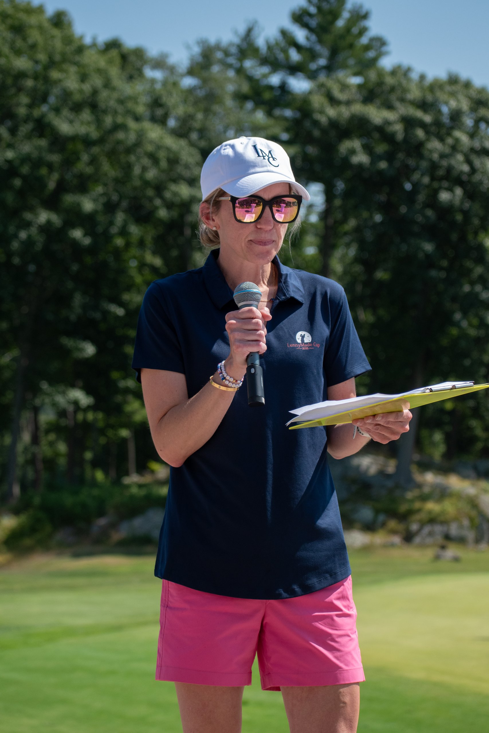 Woman in a navy LennyMade Cup polo, white LMC cap and pink shorts addressing the crowd with a microphone and clipboard.