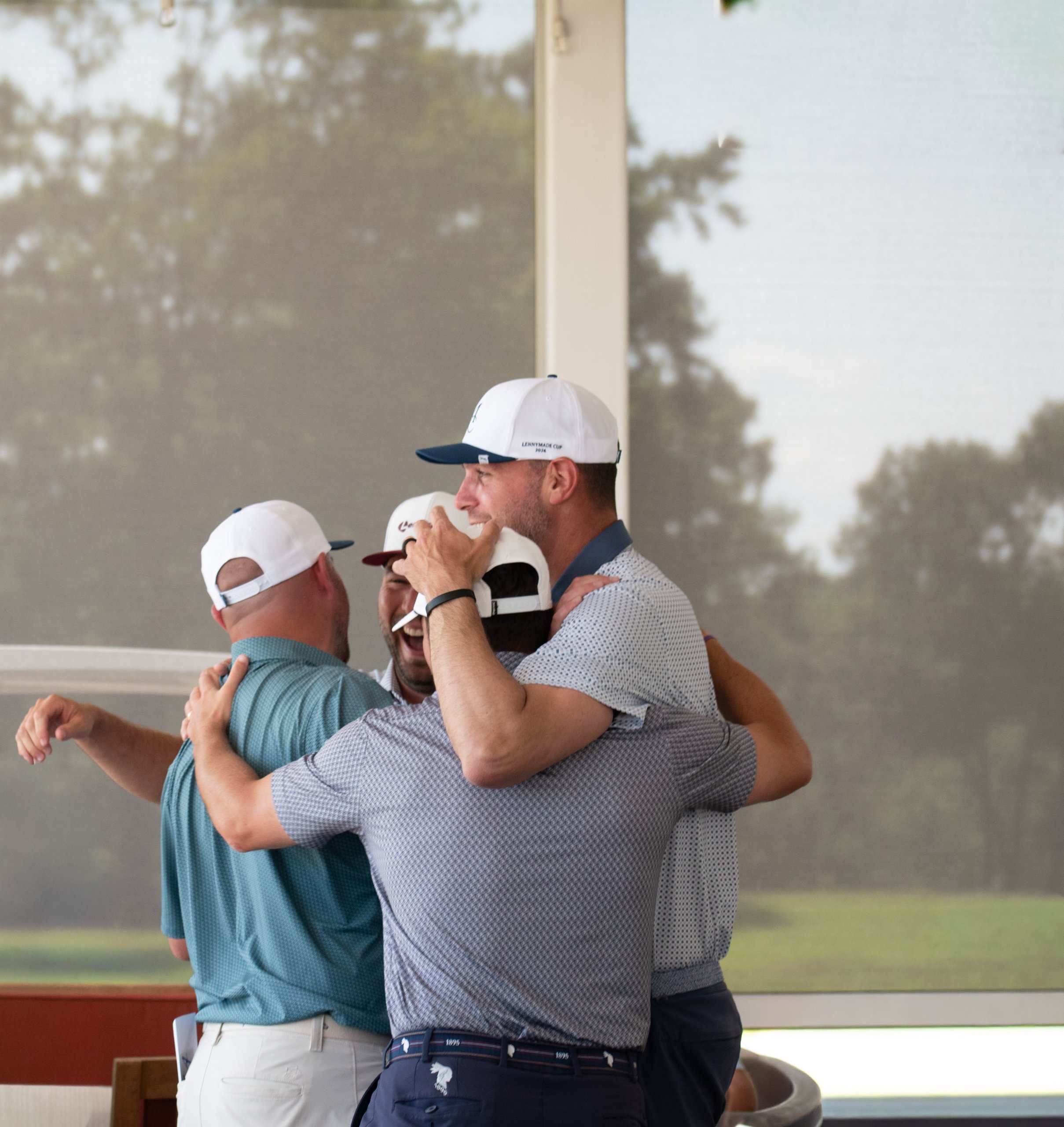 Group of golfers in a celebratory group hug on the clubhouse porch, one laughing, wearing LennyMade Cup caps.