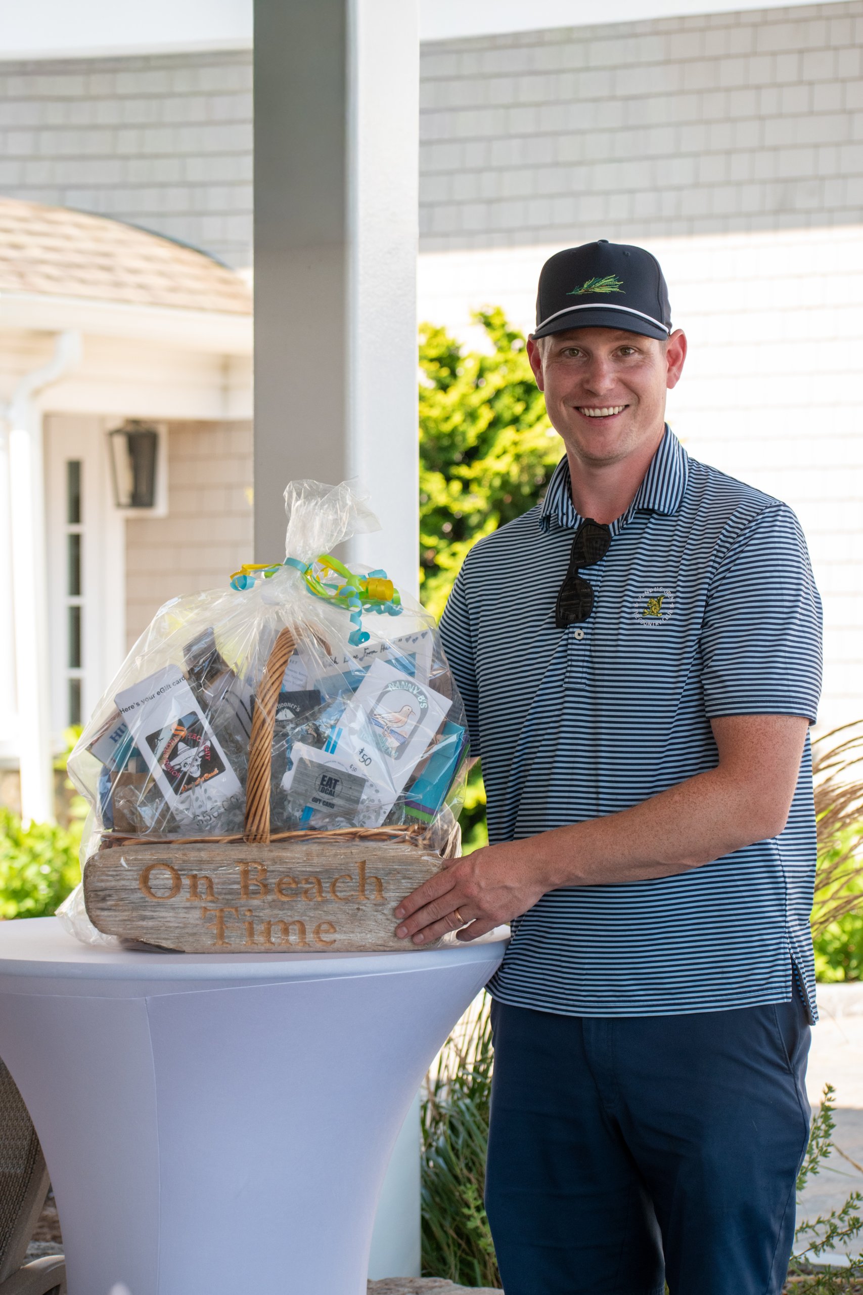 Golfer in a striped polo and navy cap smiling beside an On Beach Time raffle gift basket on a cocktail table outside the clubhouse.