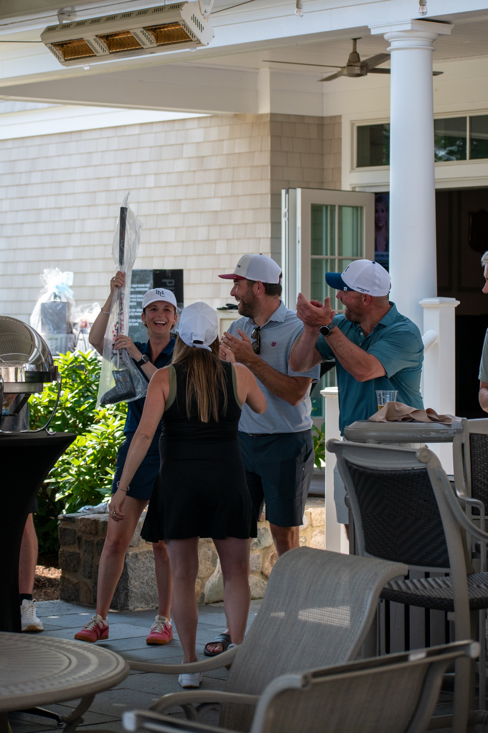 Event host in a navy LMC polo holding up a wrapped putter prize as guests applaud on the clubhouse patio.