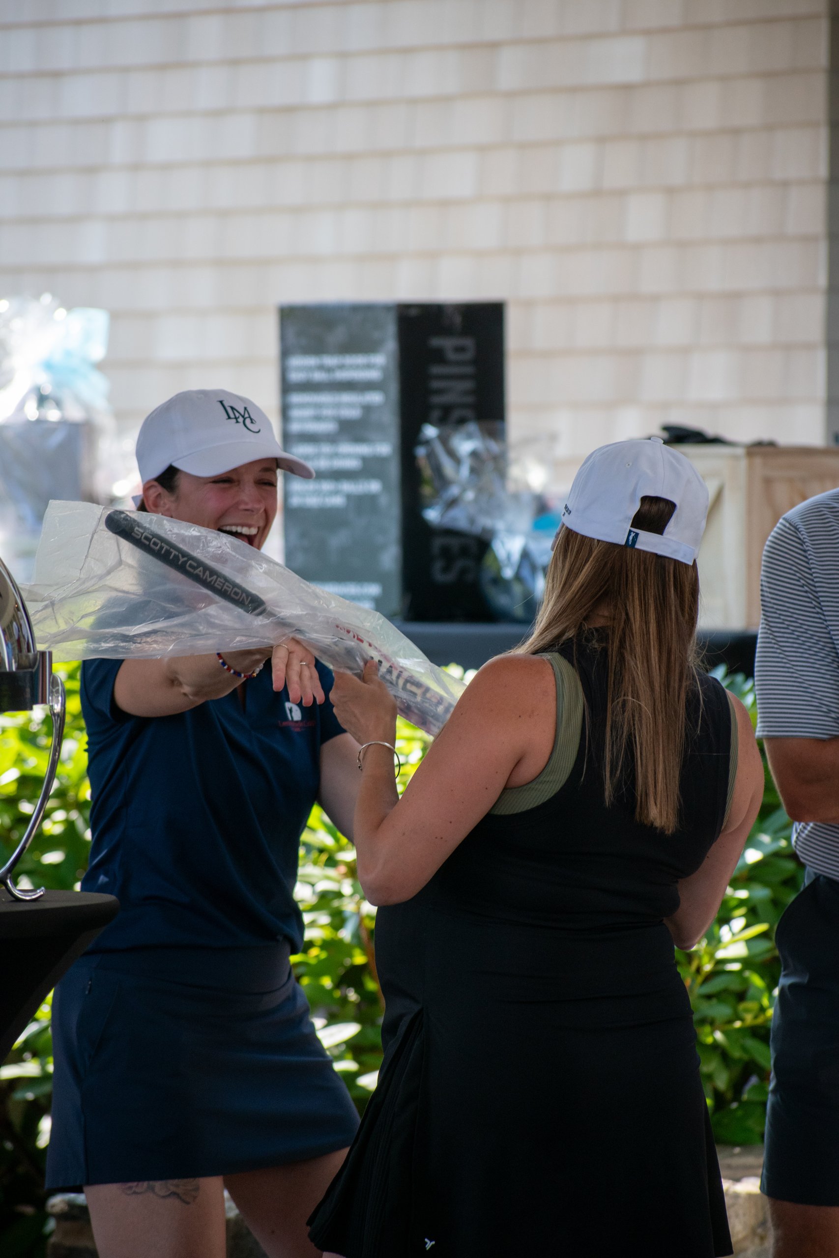 Host in an LMC cap handing a wrapped Scotty Cameron putter to a guest, both laughing.