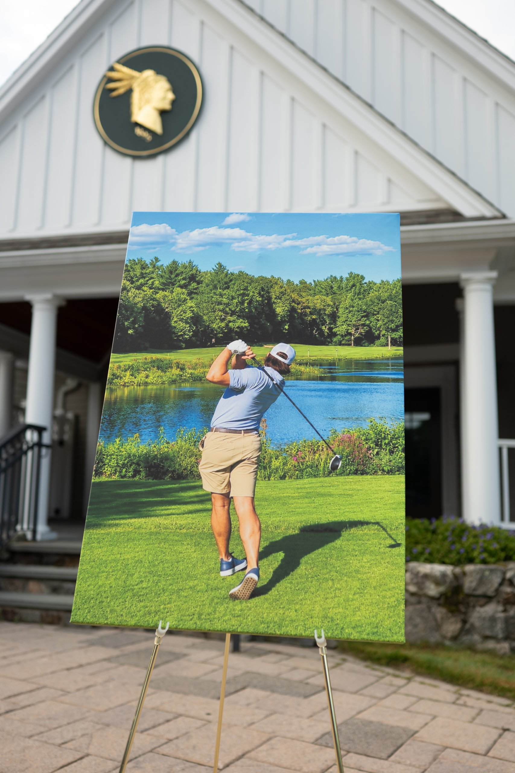 An easel holding a large canvas print of a golfer teeing off toward a pond, set in front of the clubhouse with its gold emblem.