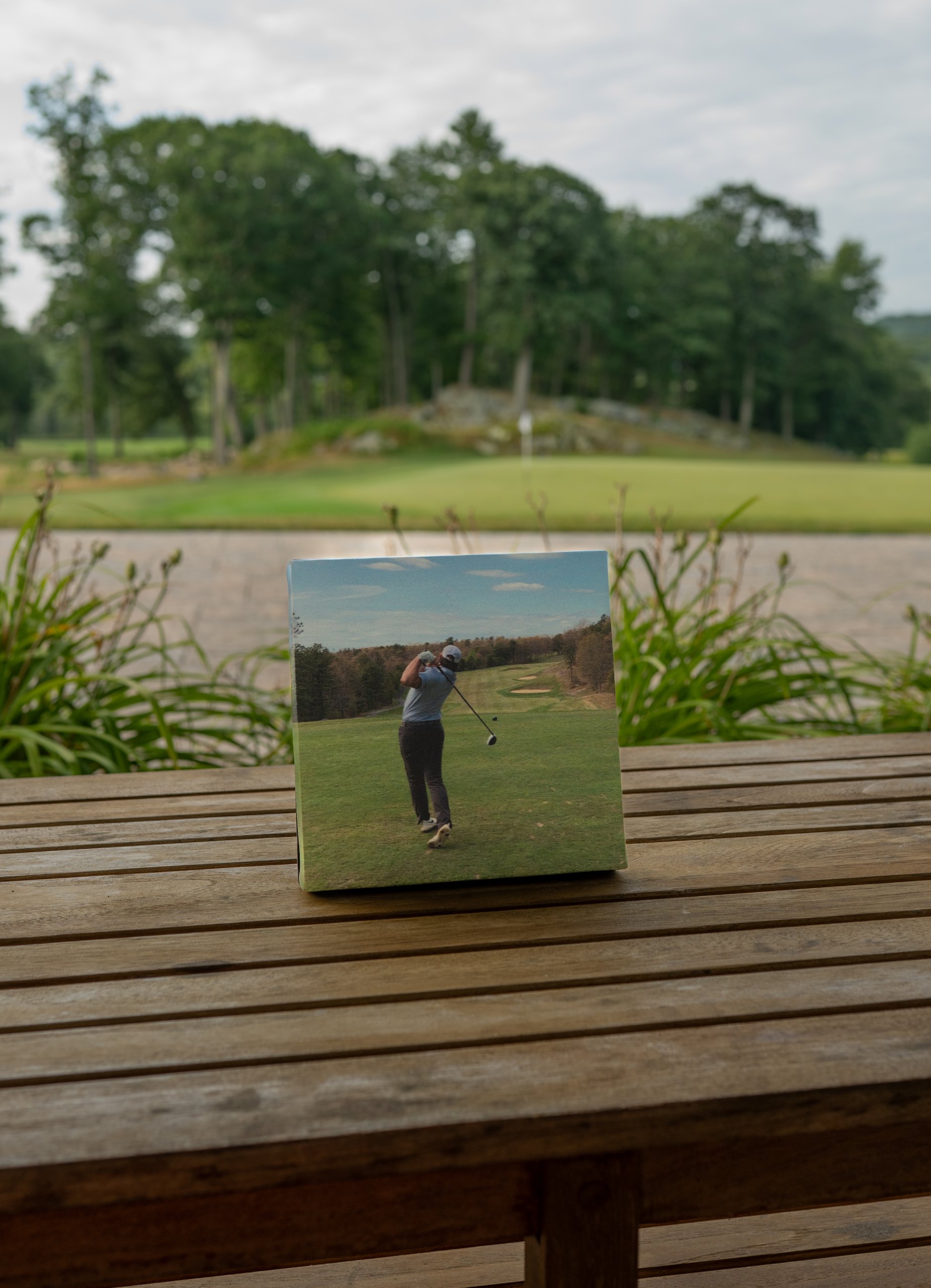 A small square canvas print of a golfer mid-swing propped on a wooden table, the course and pond blurred behind.