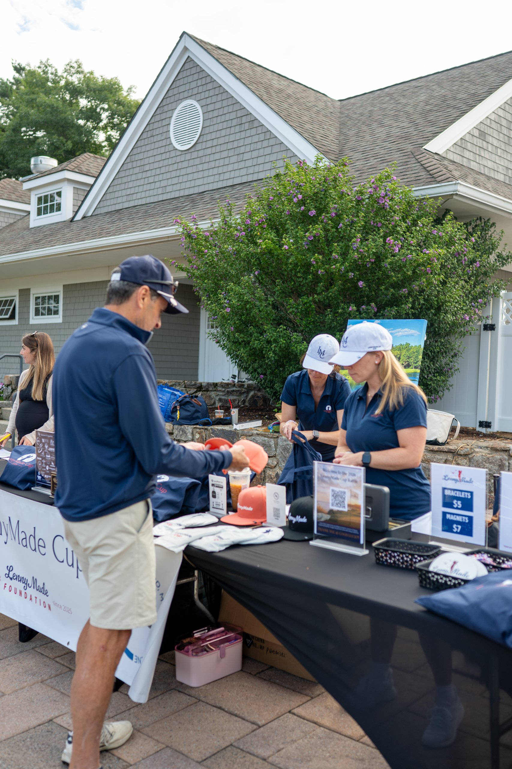 LennyMade Cup registration table with staff in LMC caps and navy polos, a flowering bush and LennyMade Foundation banner behind.