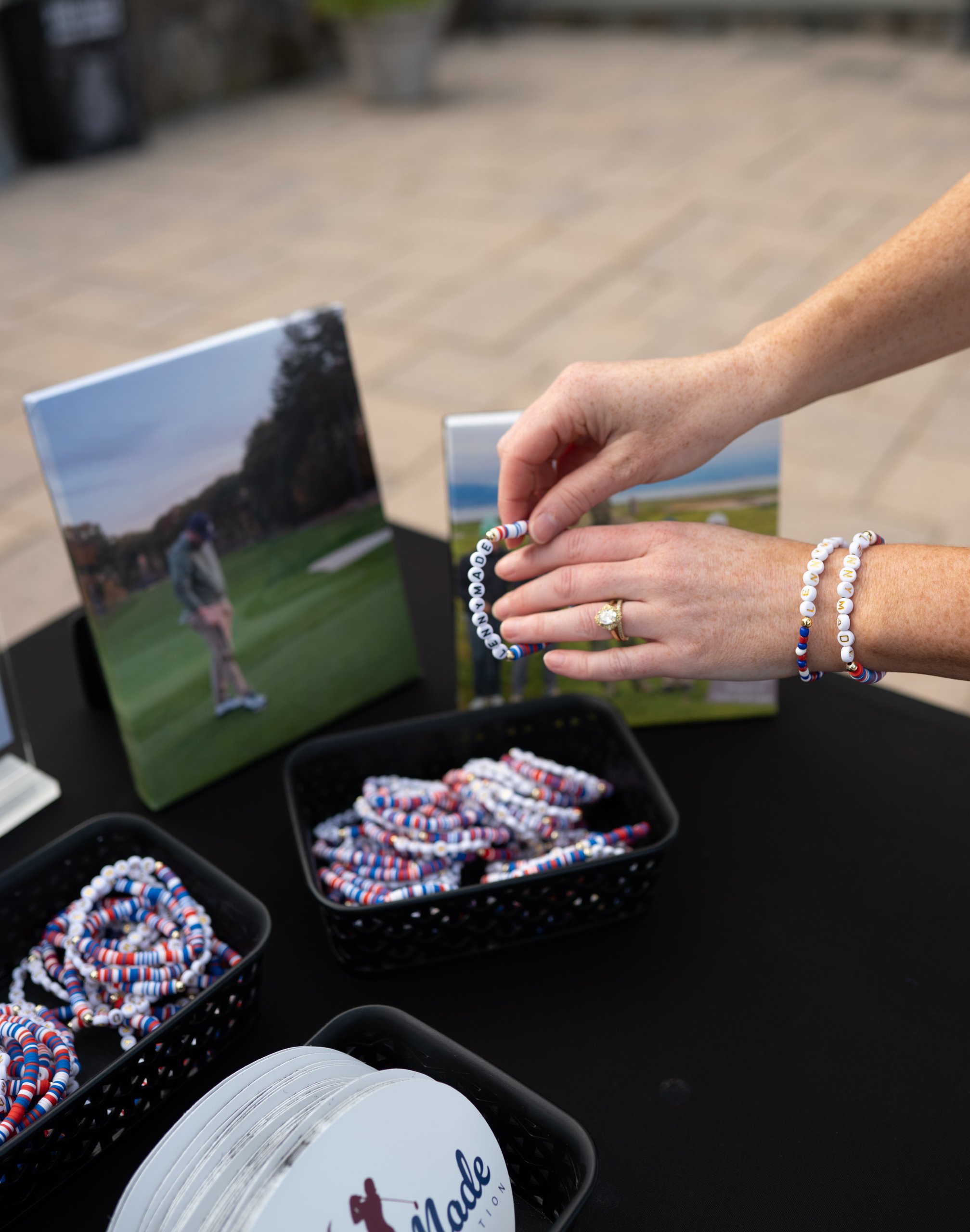 A woman's hands placing a beaded LENNYMADE bracelet beside canvas photo prints and trays of bracelets on the merch table.
