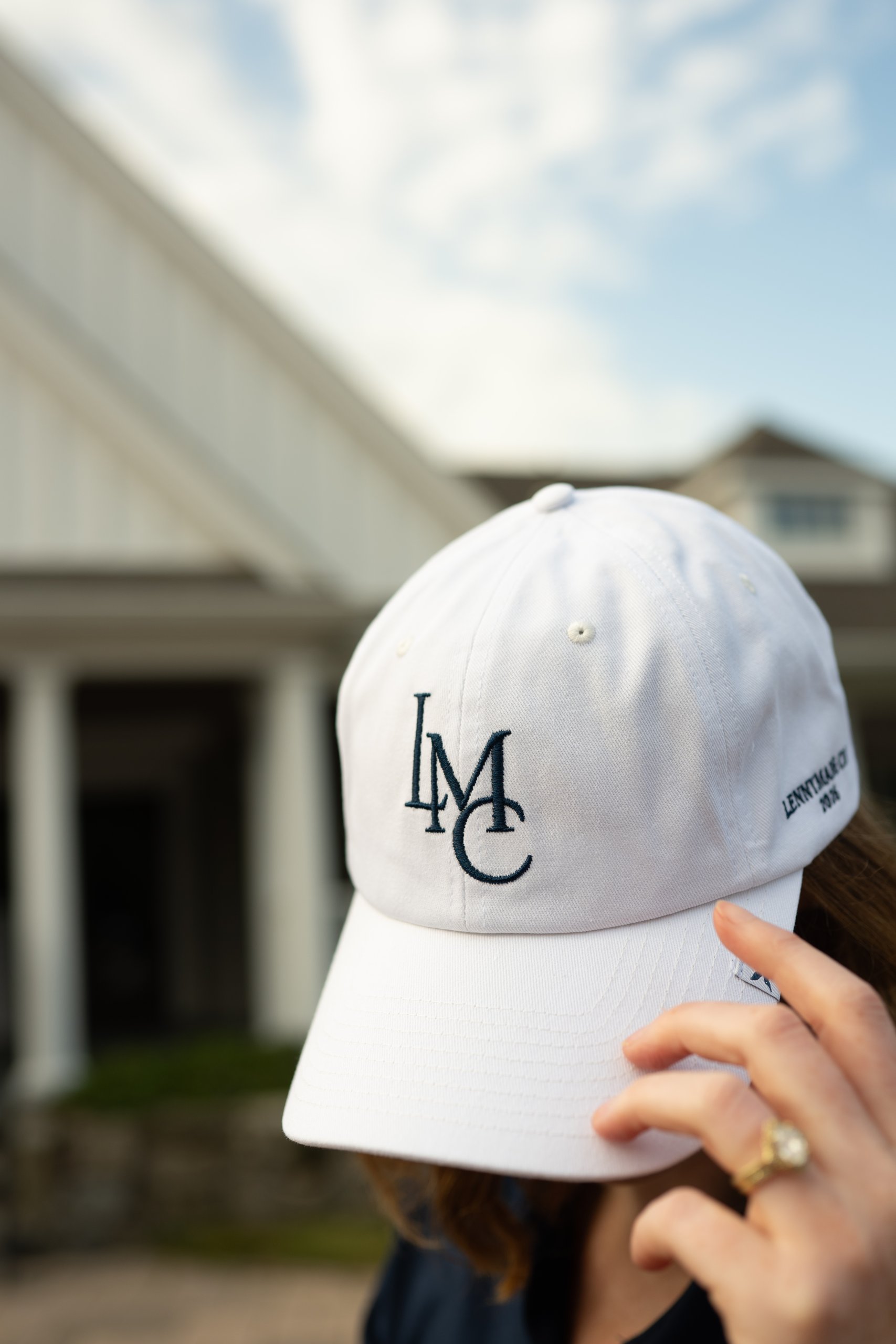 White LennyMade Cup 2026 cap with the LMC monogram held up in front of the clubhouse.