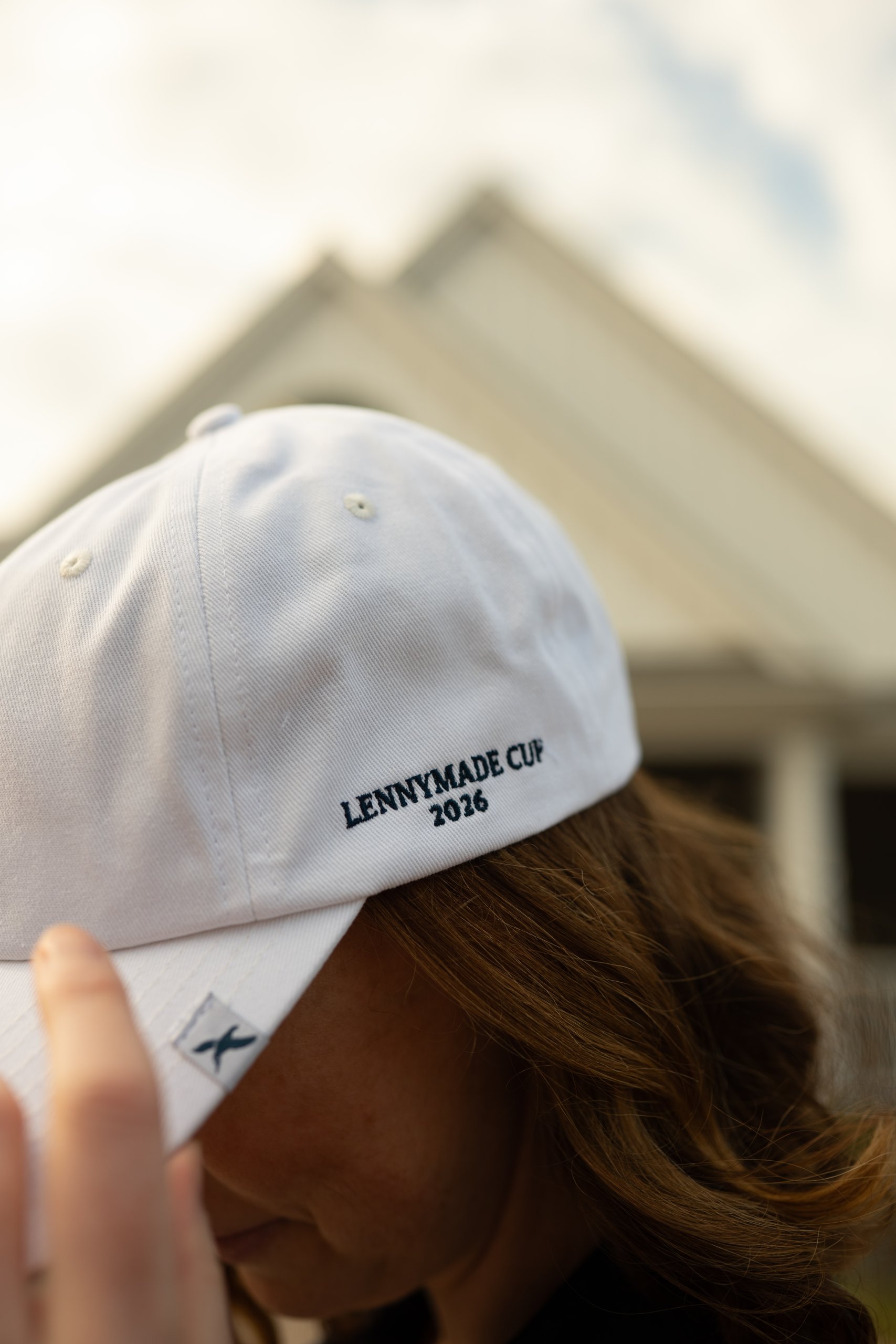 Close-up of a white cap embroidered LENNYMADE CUP 2026 held beside a woman on the clubhouse patio.