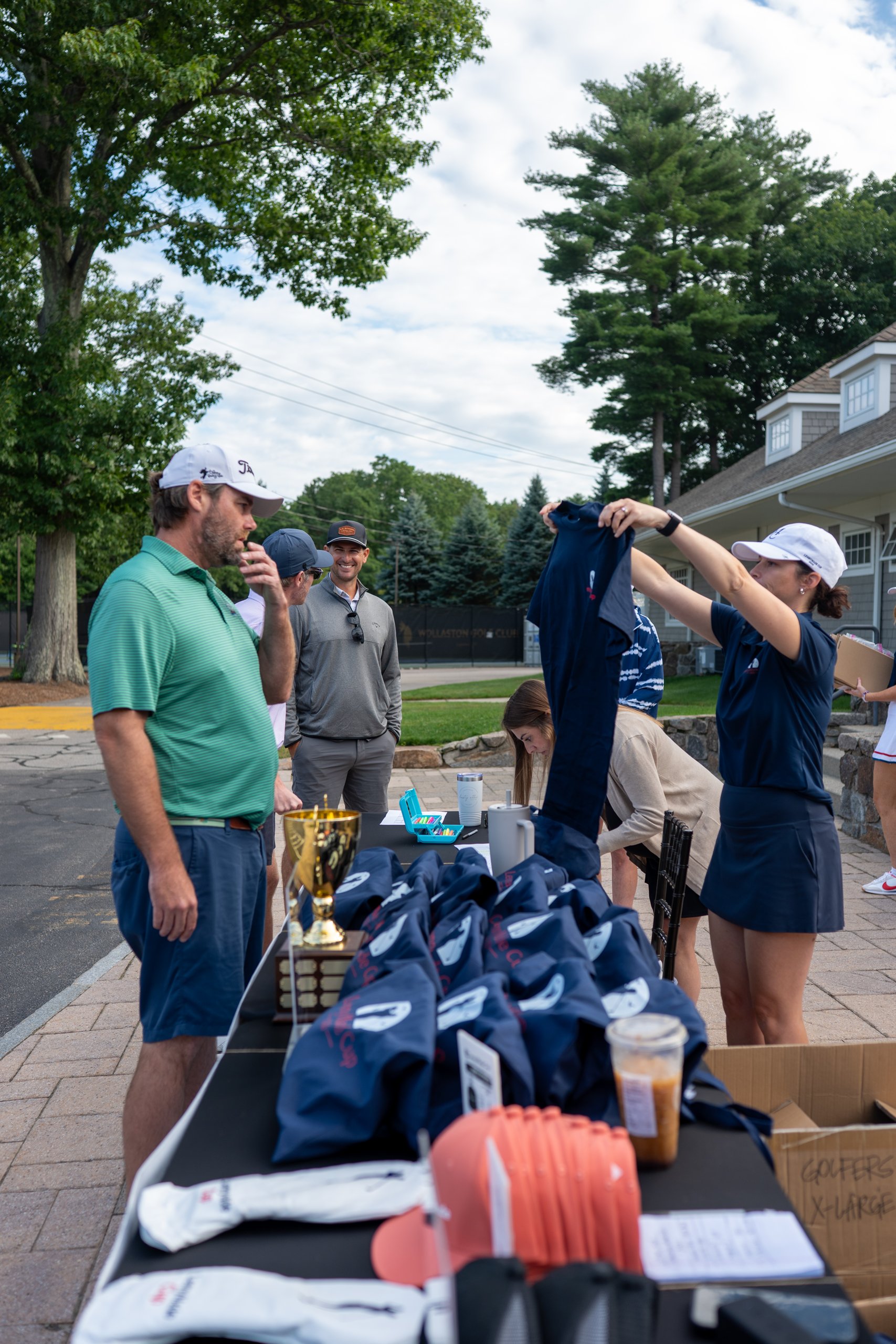 Check-in table with the perpetual LennyMade Cup trophy and navy tote bags as staff hand a golfer his tournament shirt.